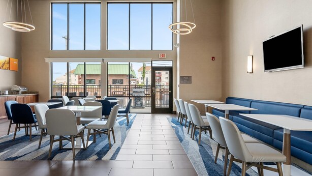 A modern dining area with tables and chairs, a blue and white striped rug, and a large window offering a view of buildings outside. - Comfort Inn Aikens Center (Martinsburg)