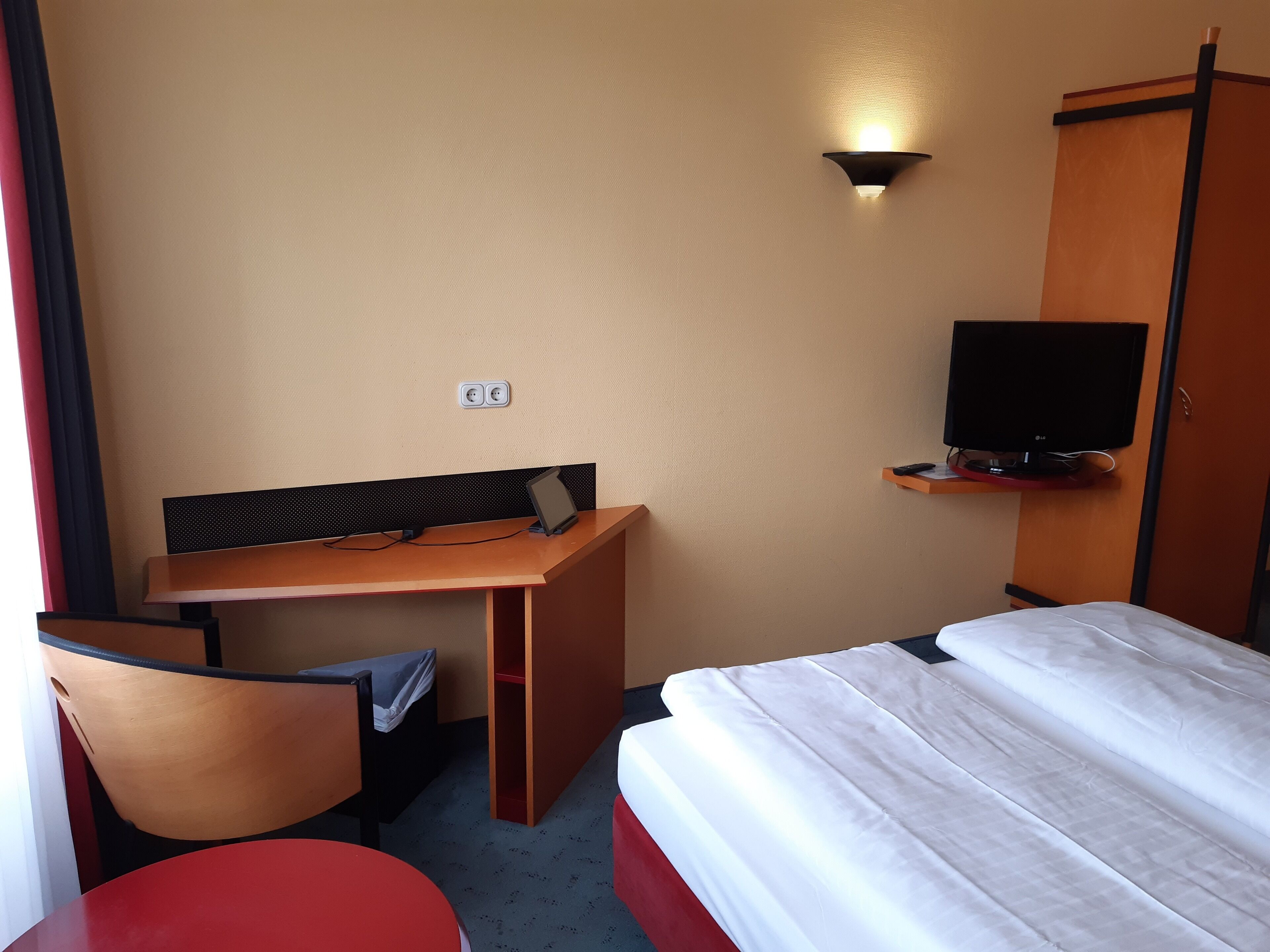 A hotel room with a bed, a desk with a chair, a television on a stand, and a wall socket.