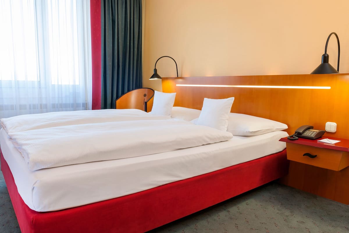 Ein Doppelbett mit weißer Bettwäsche, ein Holzkopfende, ein Nachttisch mit Lampe, Telefon und einer kleinen Schublade.