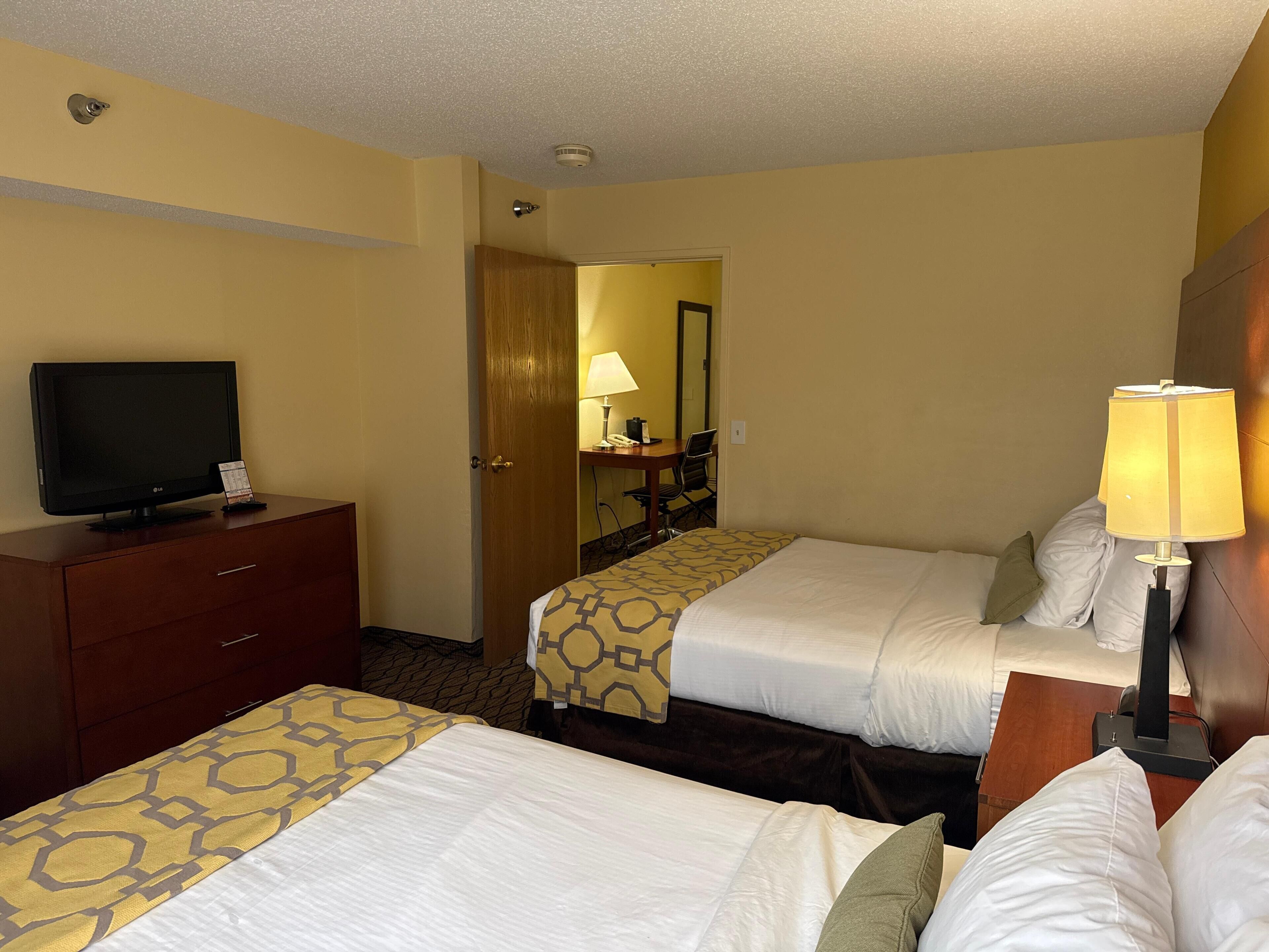 A hotel room with two beds, a TV on a dresser, a desk with a lamp, and a mirror on the wall.