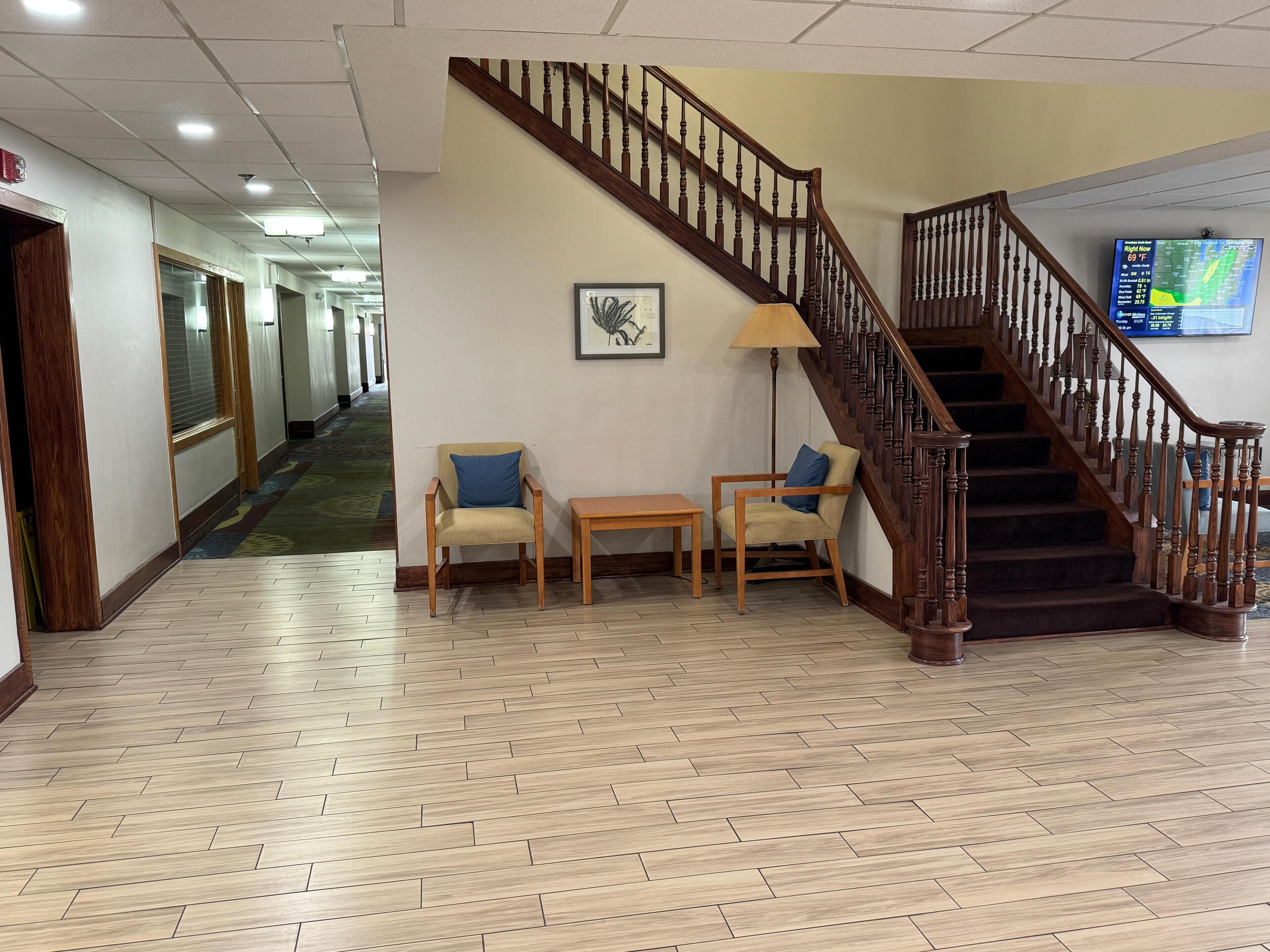 Recepció d'hotel amb una escala de fusta, dues cadires amb coixins, una taula petita i un quadre encimbellat a la paret.