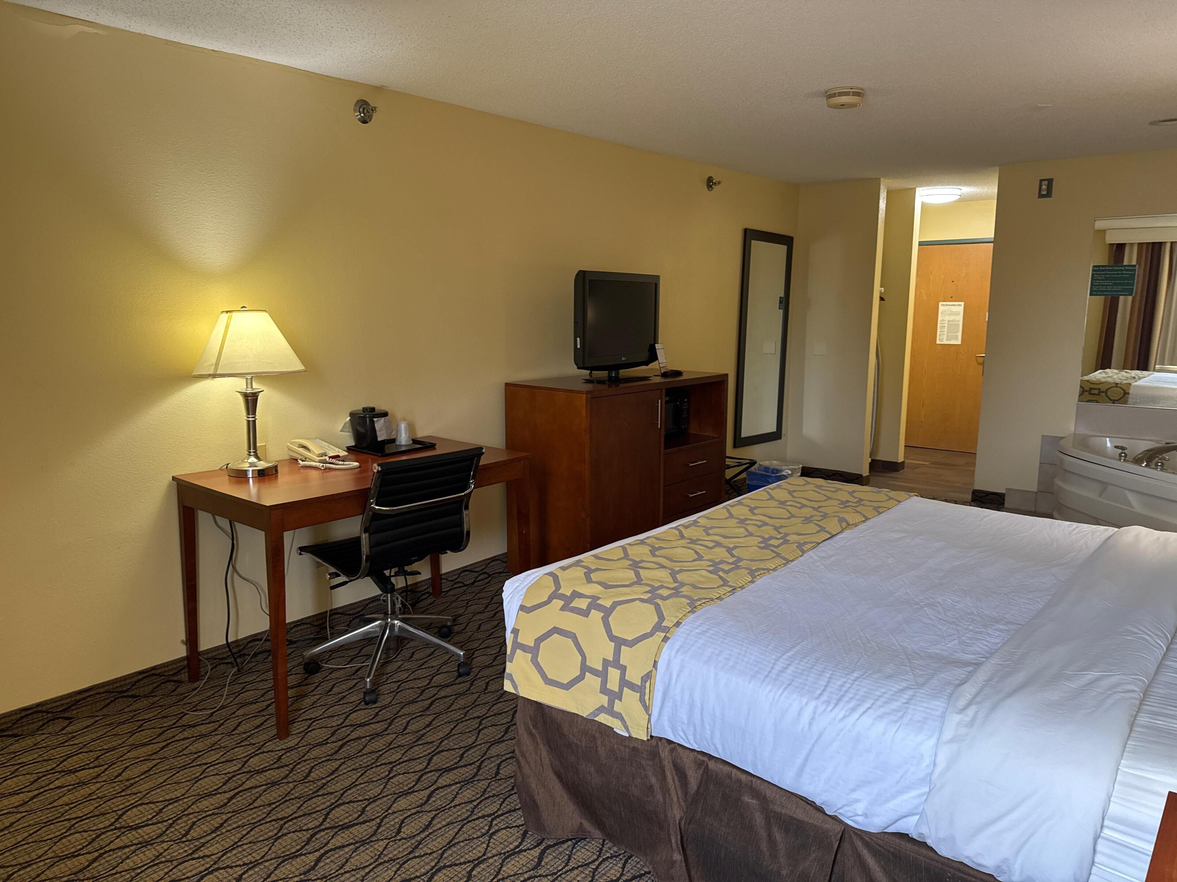 Habitación de hotel con cama, escritorio, silla, lámpara, televisión y baño con lavamanos y espejo.
