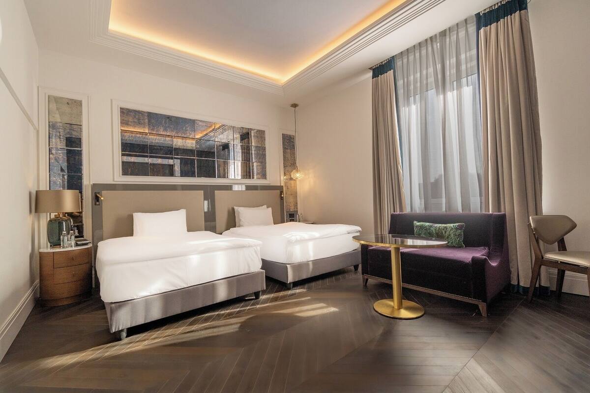 A hotel room with two beds, a sofa, a small table, and a large window with curtains.