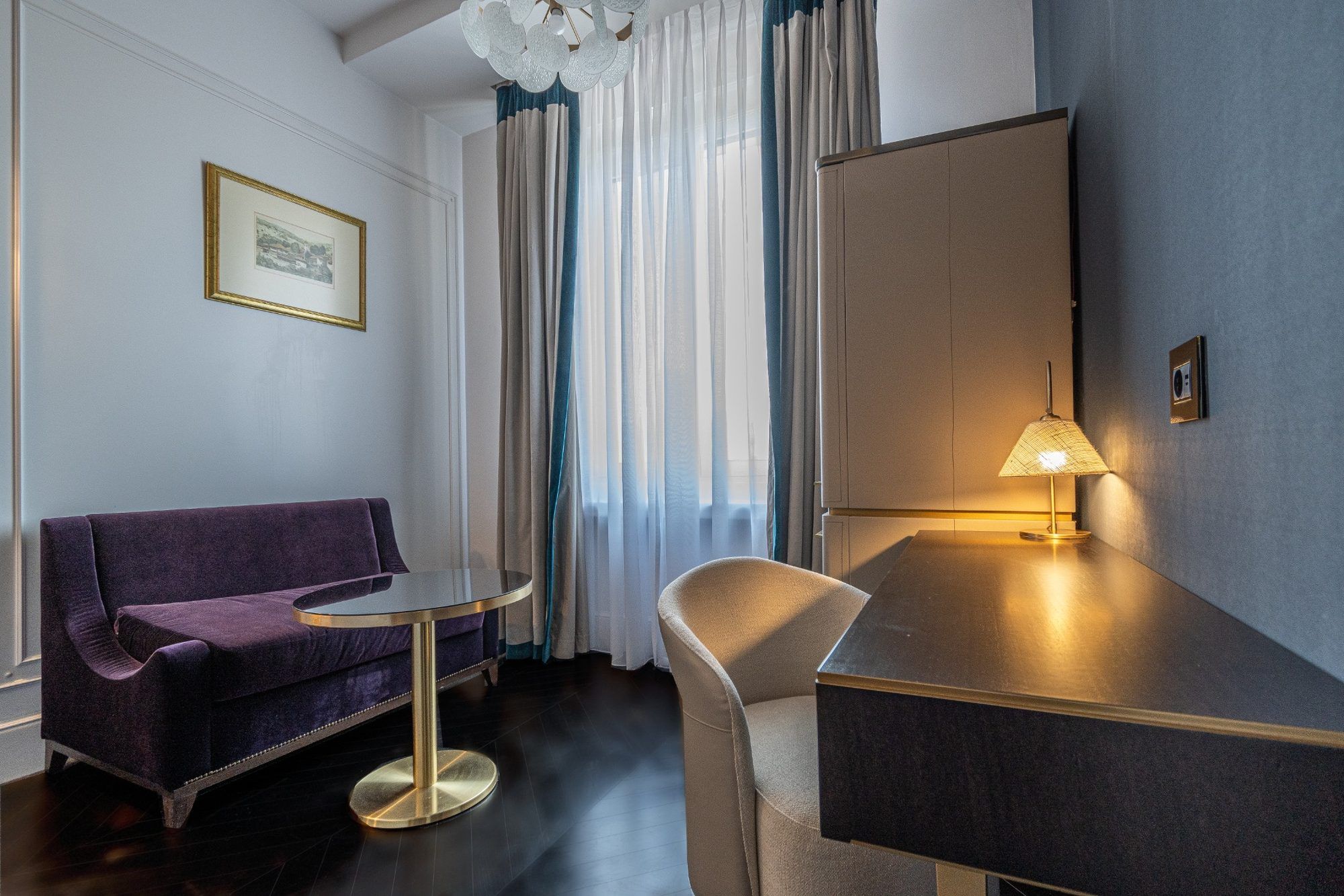 A hotel room with a purple sofa, a small round table, a lamp, a large mirror, and a framed picture on the wall.