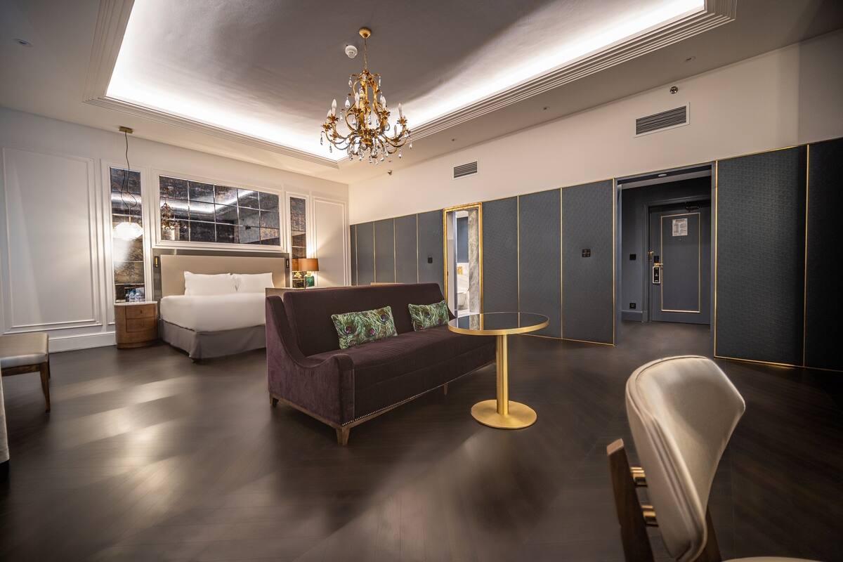 A hotel room with a bed, a sofa, a round table, and a chair. There is a chandelier above the sofa.