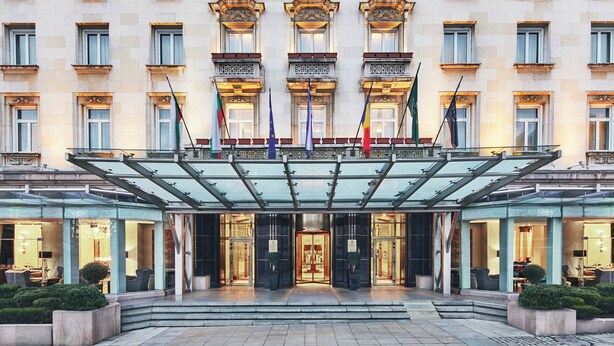 A hotel entrance with a glass canopy, multiple flags, and a well-lit facade. - Sofia Balkan Palace (Sofia)