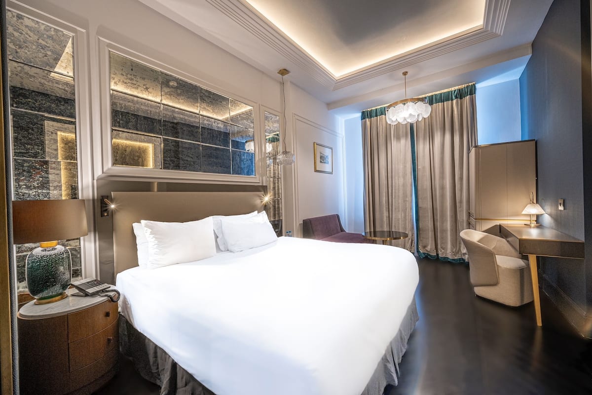 A modern hotel room with a large bed, a desk, a chair, a phone, and a window with a view.