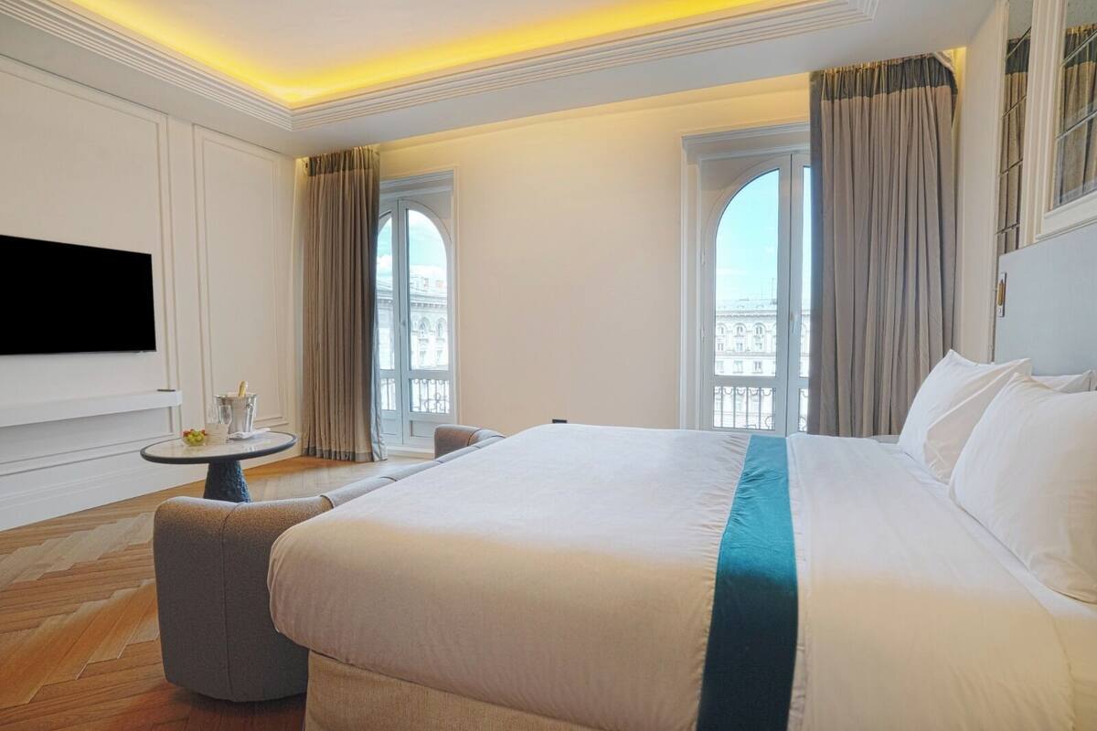 A hotel room with a large bed, a TV, a small table with a drink, and a view of a building through the window.