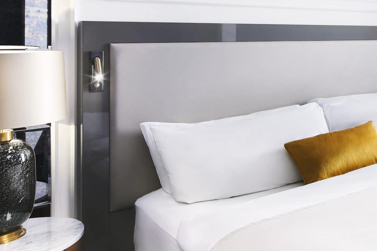 A neatly made bed with white linens and a mustard-colored pillow, a bedside lamp, and a wooden nightstand.