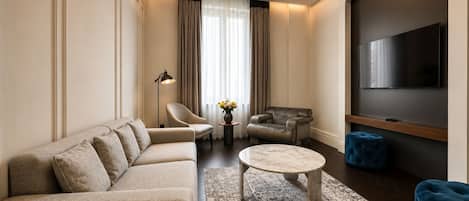 A modern living room with a sectional sofa, a round coffee table, a blue ottoman, a leather armchair, a small side table with a vase, and a flat-screen TV mounted on the wall.