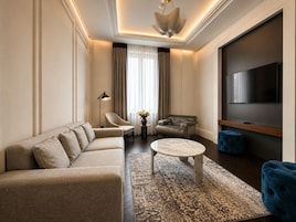 Ein modernes Wohnzimmer mit einem Ecksofa, einem runden Couchtisch, einem blauen Hocker, einem Ledersessel, einem kleinen Beistelltisch mit einer Vase und einem flachbildfernseher an der Wand.