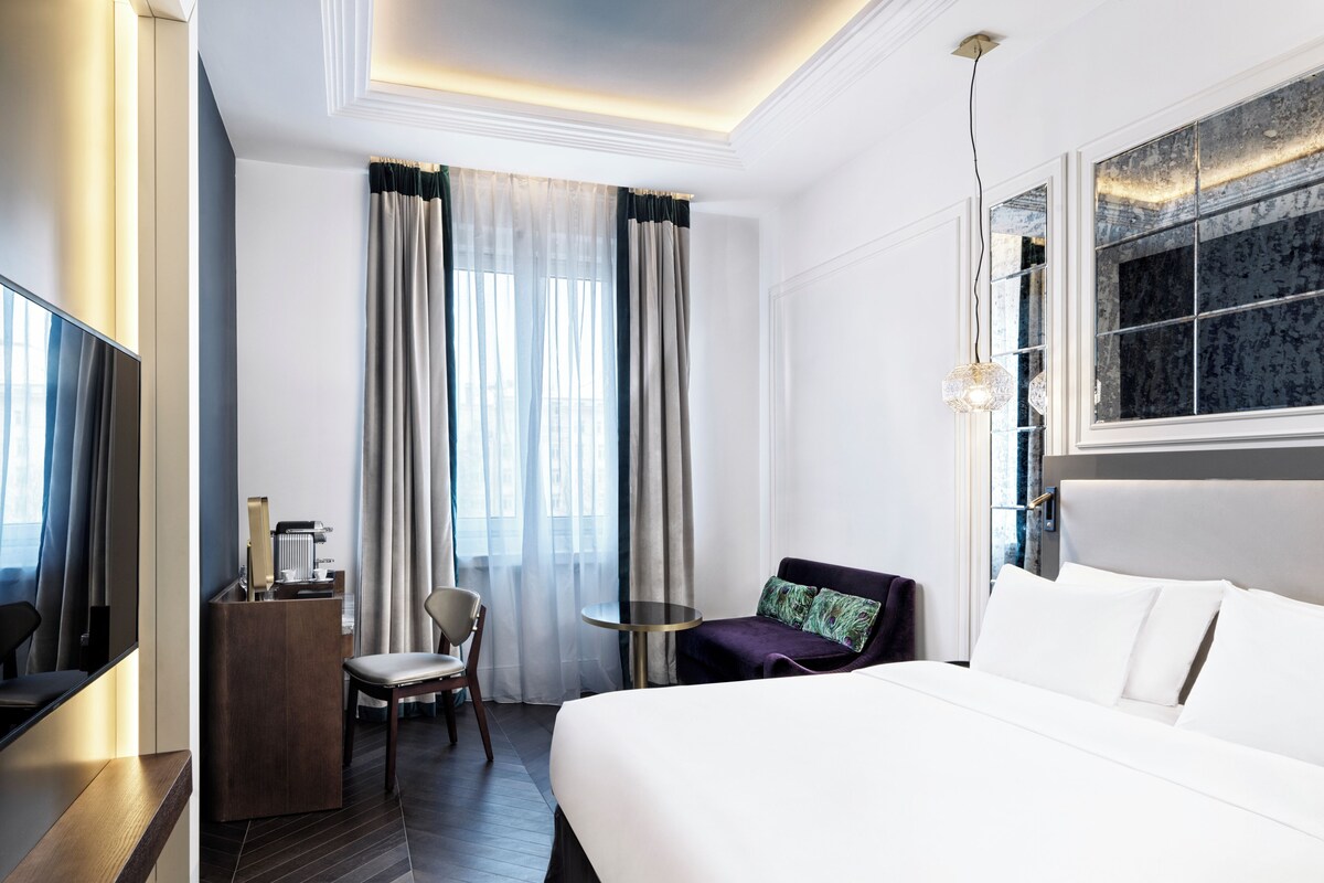 A hotel room with a bed, a desk, a chair, a small table, and a TV.