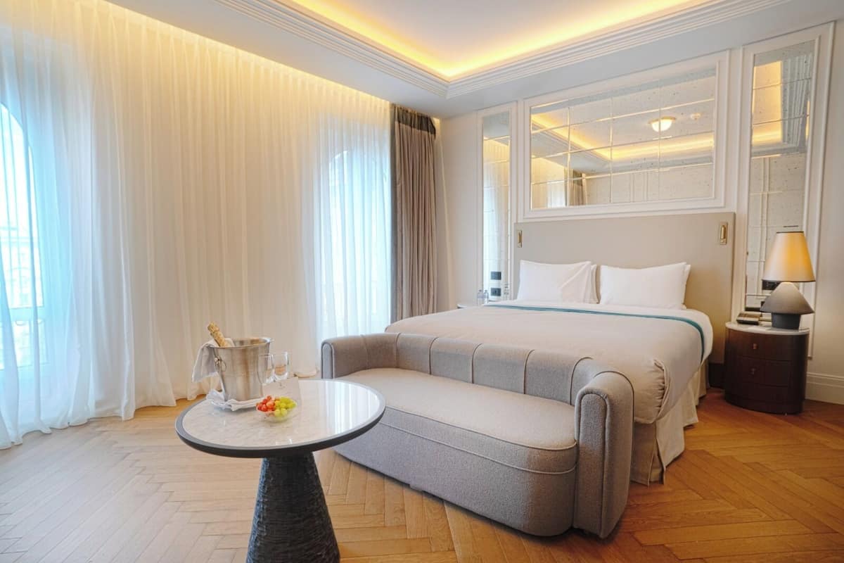 A hotel room with a bed, a bedside table with a lamp, a small round table with a fruit bowl, and a large window with curtains.