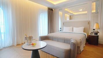 Una habitación de hotel con cama, mesita de noche con lámpara, una pequeña mesa redonda con frutero y un ventanal con cortinas.