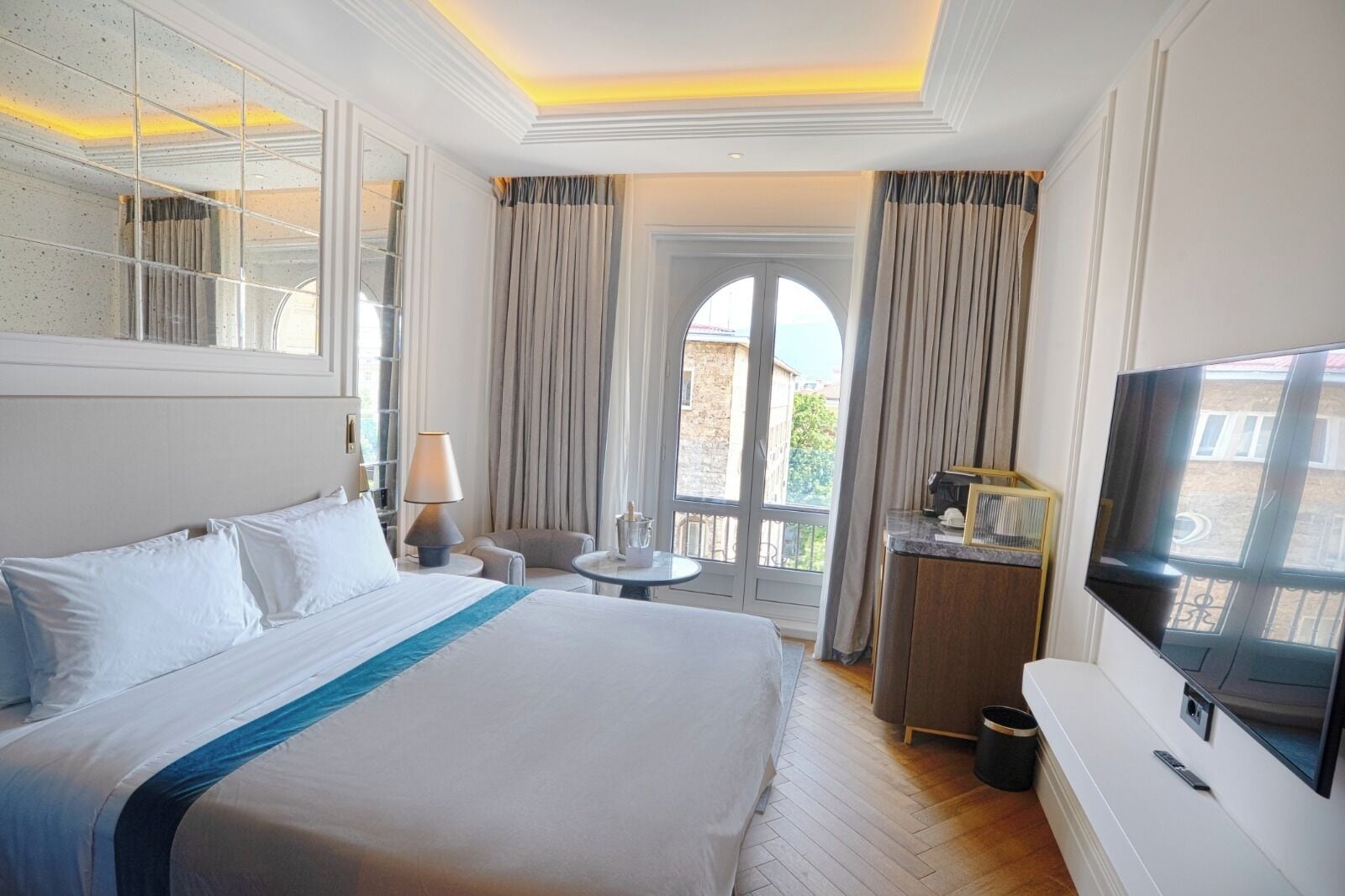 Habitación de hotel con una cama grande, un escritorio con televisor, una mesita de noche con lámpara y vistas al exterior.