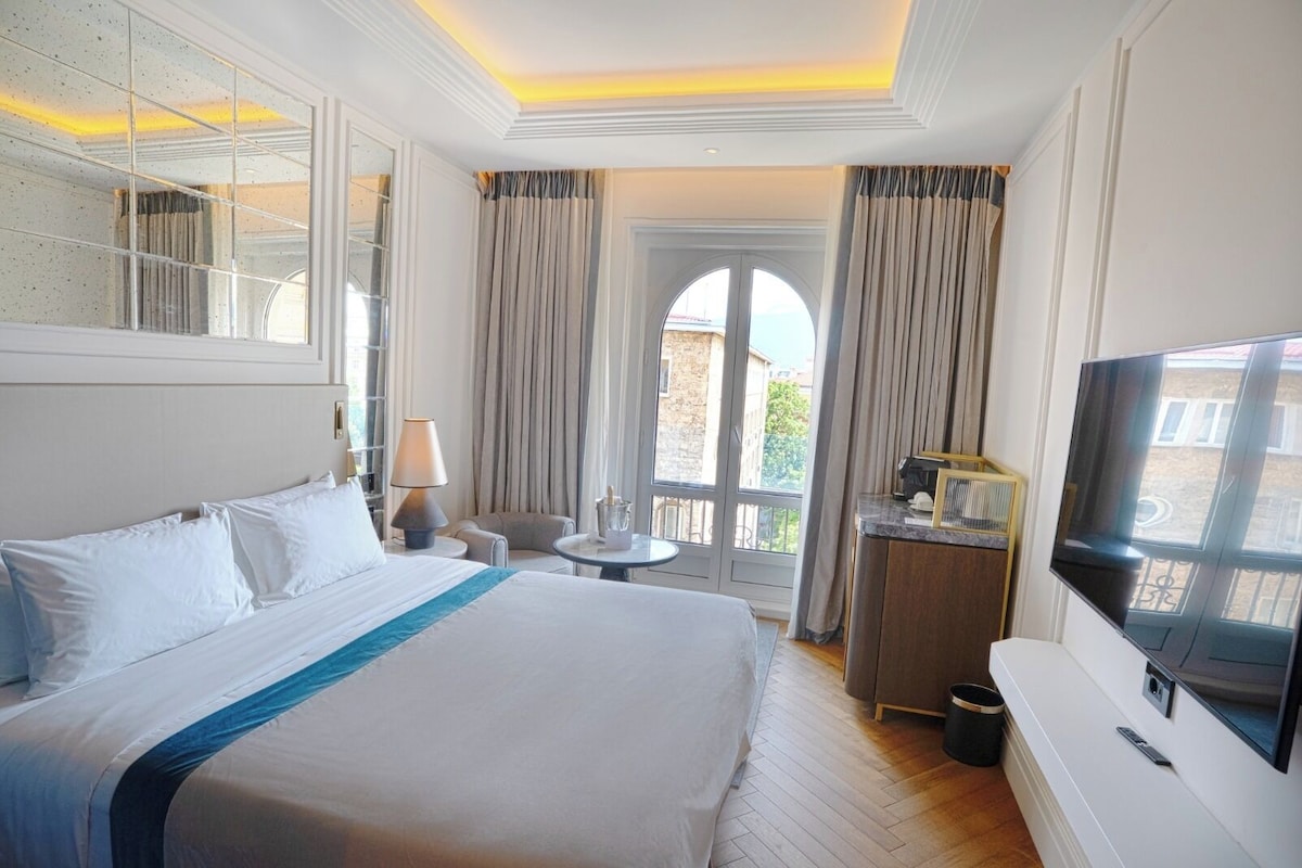 A hotel room with a large bed, a desk with a TV, a small table with a lamp, and a view of the outside.