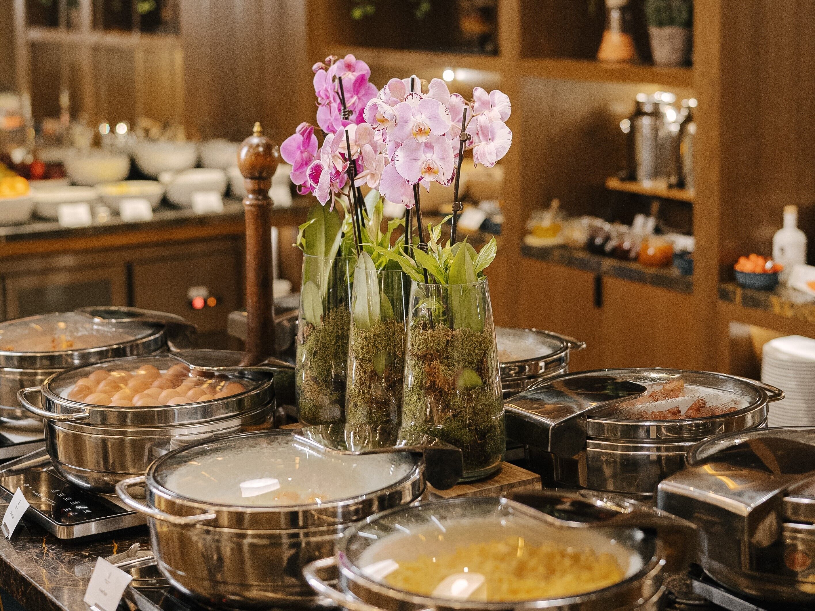 A buffet area with various food containers and a decorative vase with pink orchids.