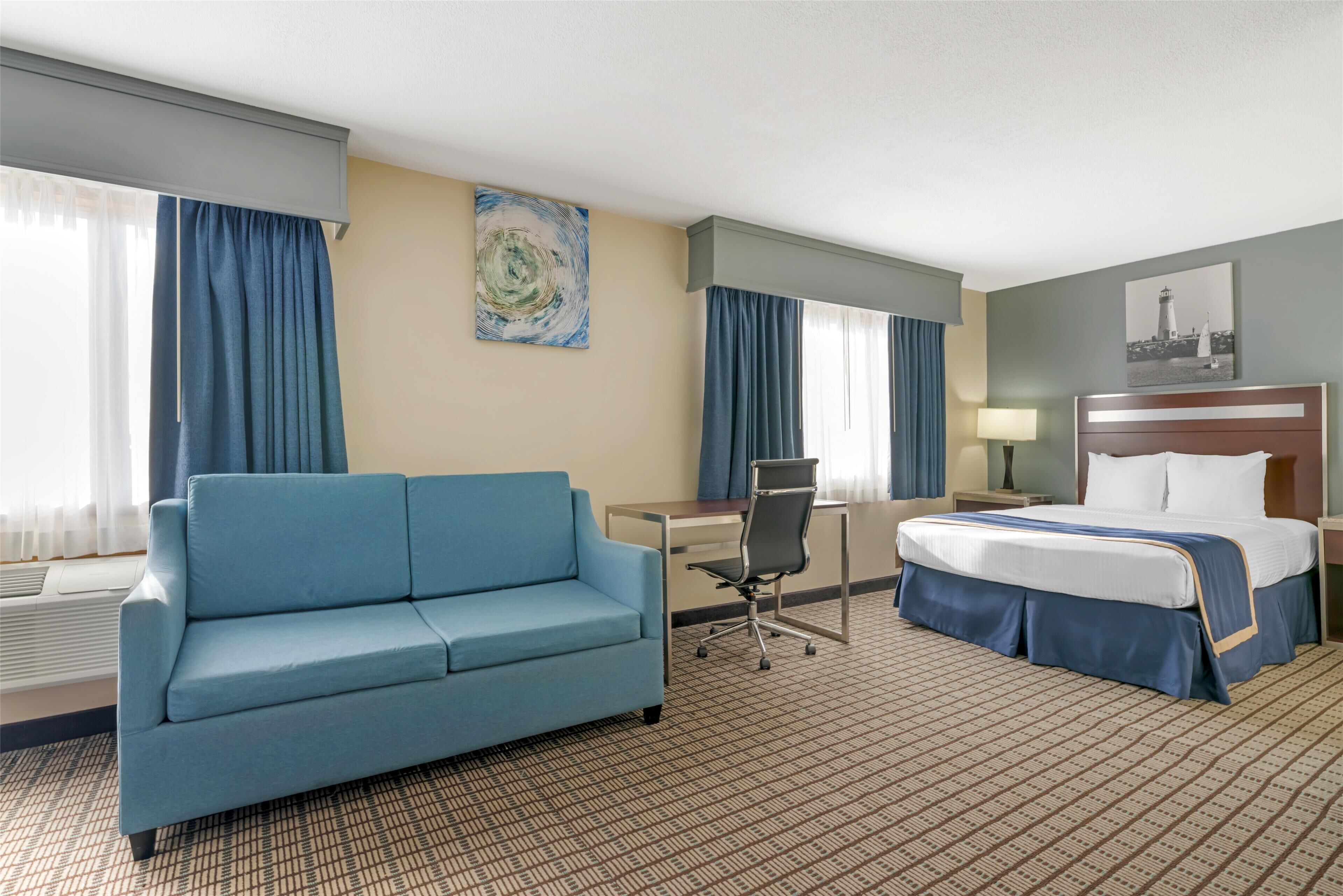 A hotel room with a blue sofa, a desk, a chair, a bed, and a nightstand with a lamp.