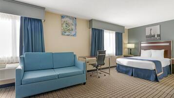A hotel room with a blue sofa, a desk, a chair, a bed, and a nightstand with a lamp.