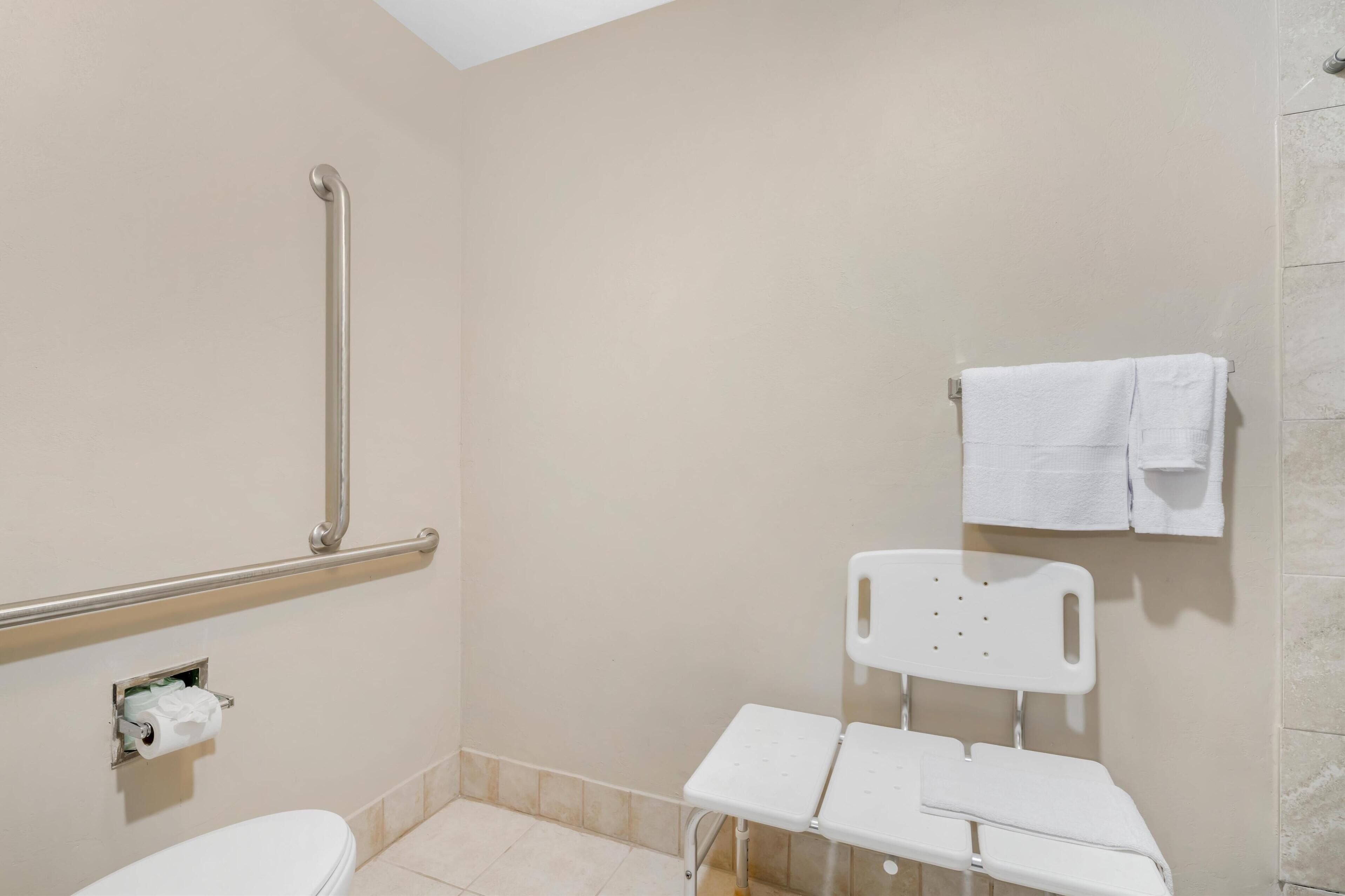 Une salle de bain équipée de toilettes, d’une barre d’appui, d’un banc blanc et d’un porte-serviettes.
