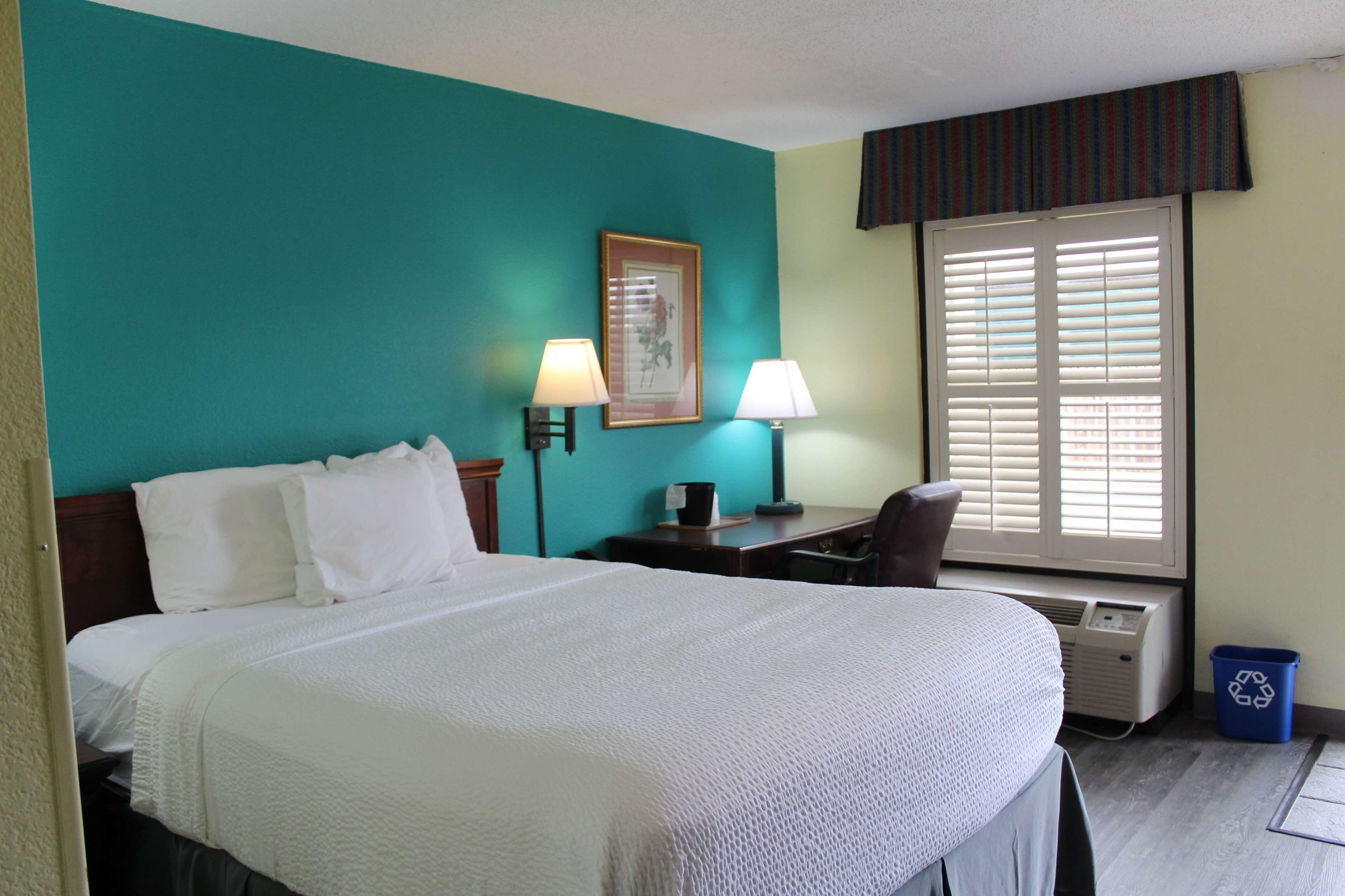 A hotel room with a large bed, two bedside lamps, a desk with a chair, a window with blinds, and a recycling bin.