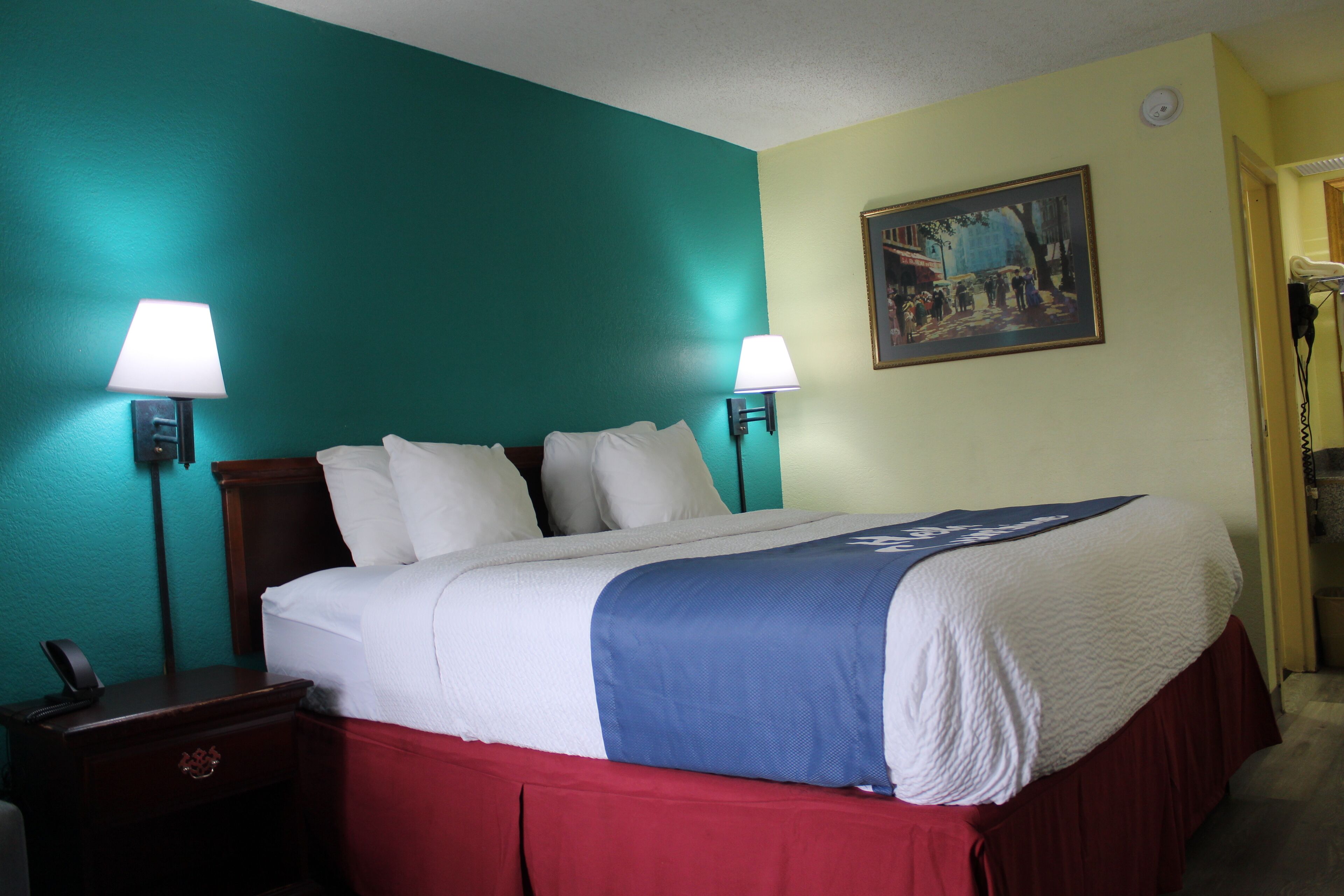 A hotel room with a bed, two bedside lamps, a nightstand, a painting on the wall, and a door leading to another room.