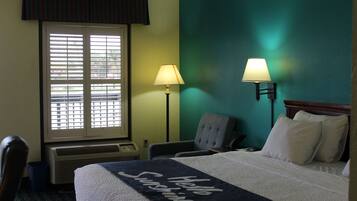Una habitación de hotel con cama, dos lámparas, una silla, ventana con persianas y un calentador de pared.