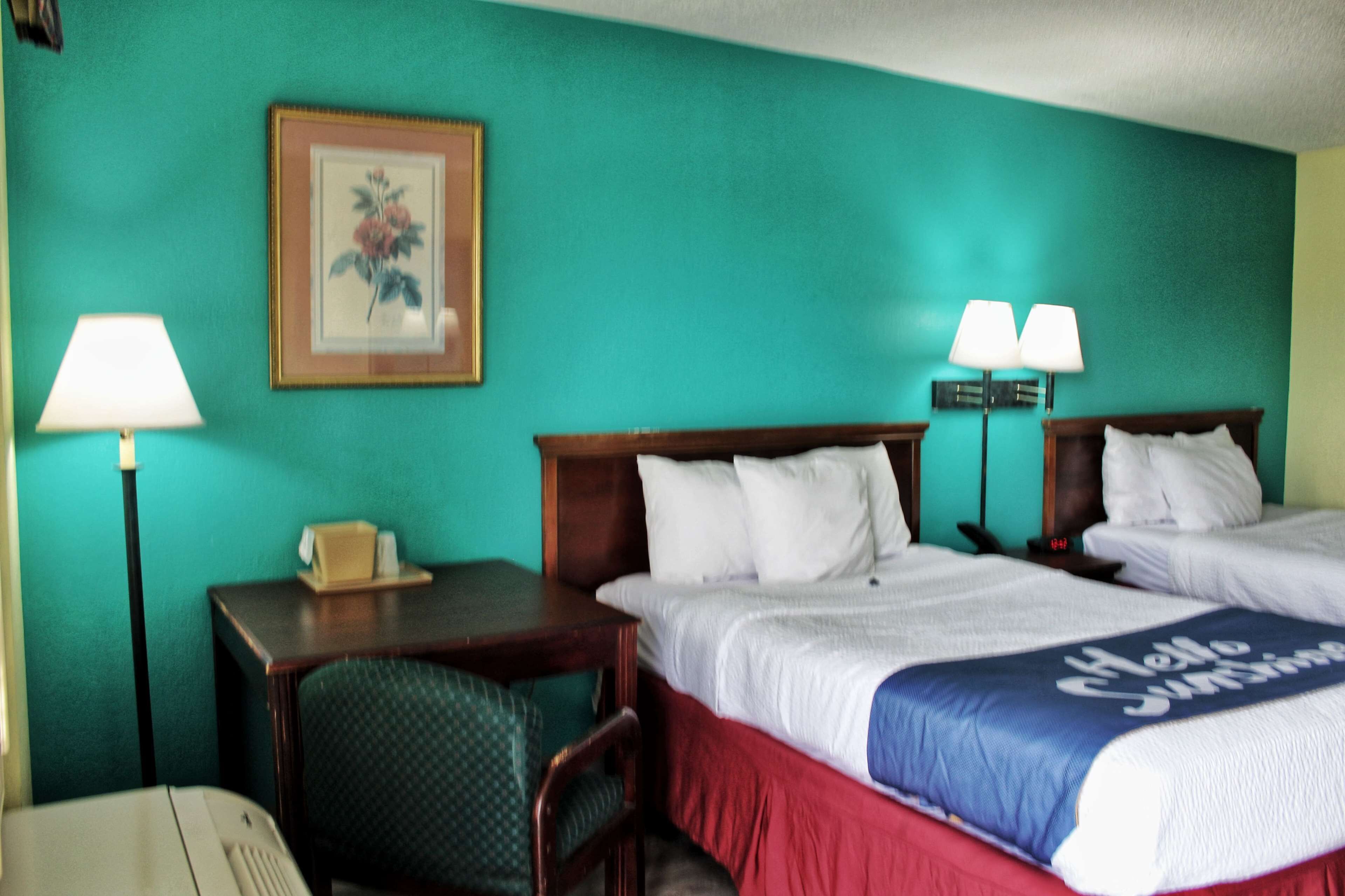 A hotel room with two beds, a desk, a chair, a lamp, and a framed picture on the wall.