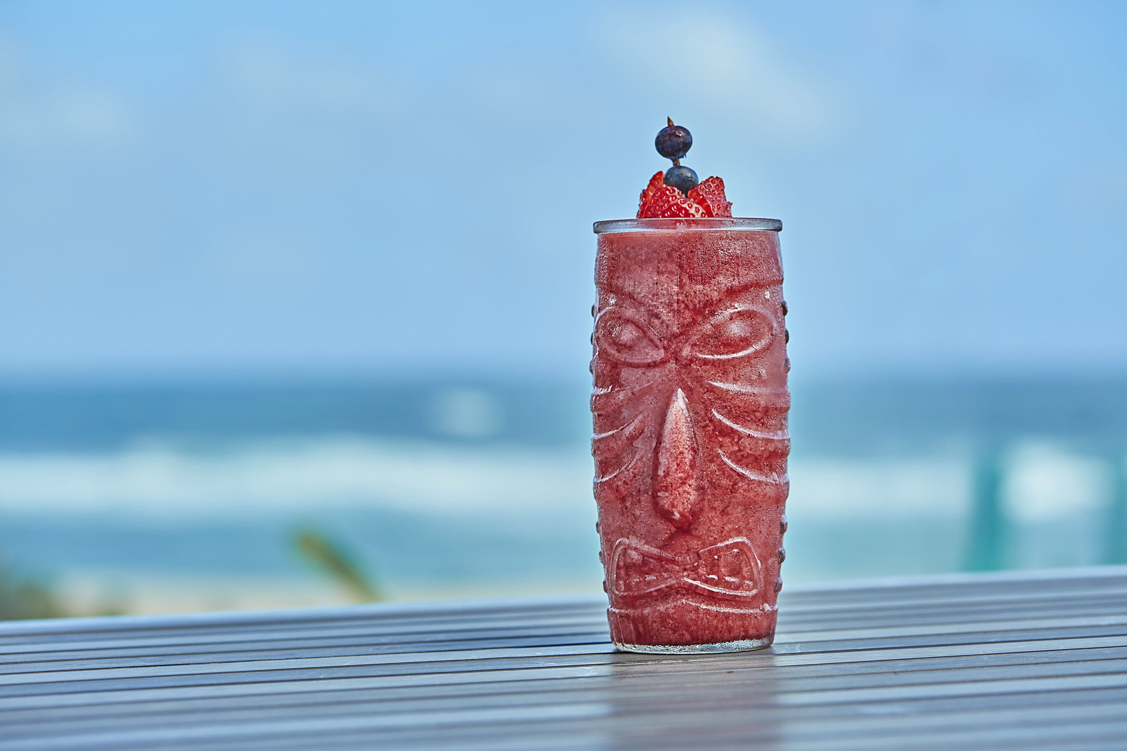 A glass with a textured design and a berry garnish on a metal surface with a blurred ocean and sky in the background.