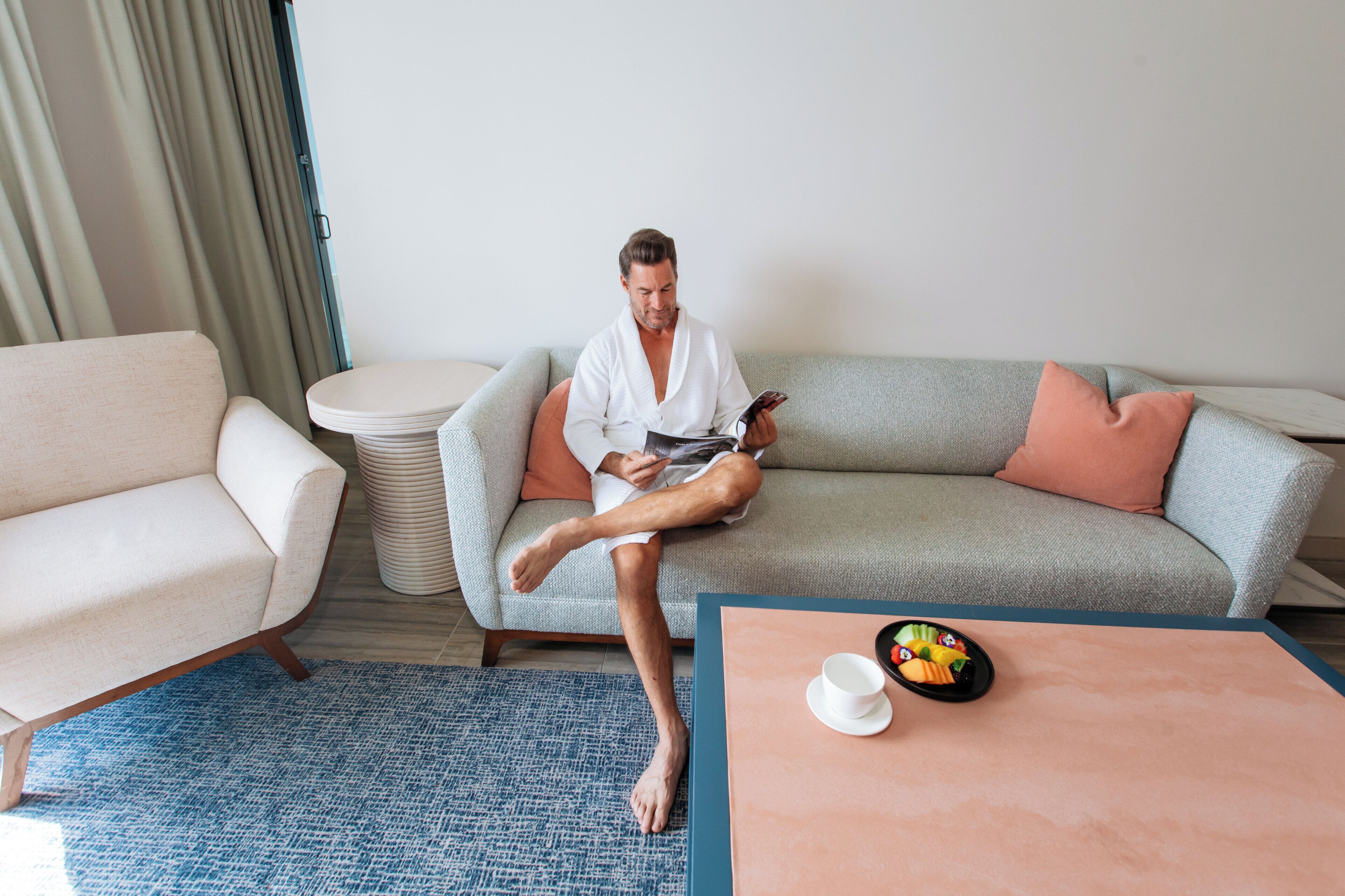 Um homem sentado em um sofá, com um smartphone, uma xícara de café e uma tigela de frutas sobre uma mesa.