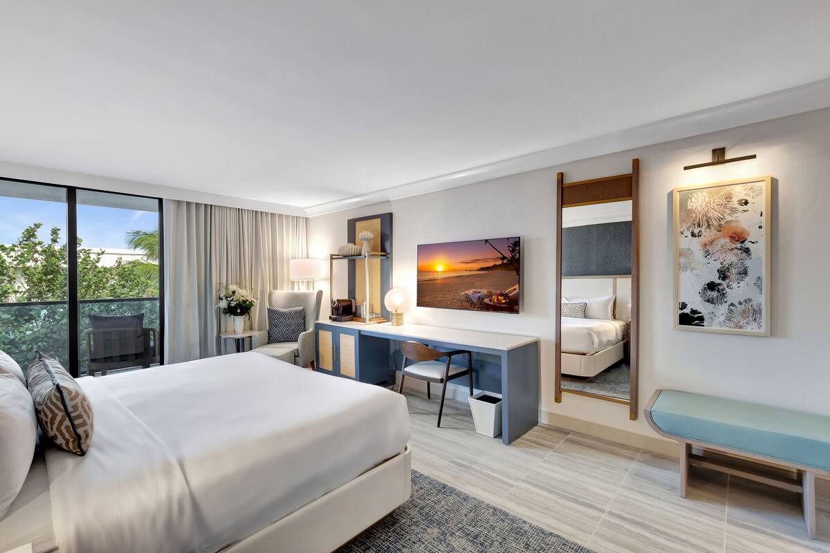 A modern hotel room with a large bed, a desk with a chair, a bench, and a large window offering a view of the outdoors.