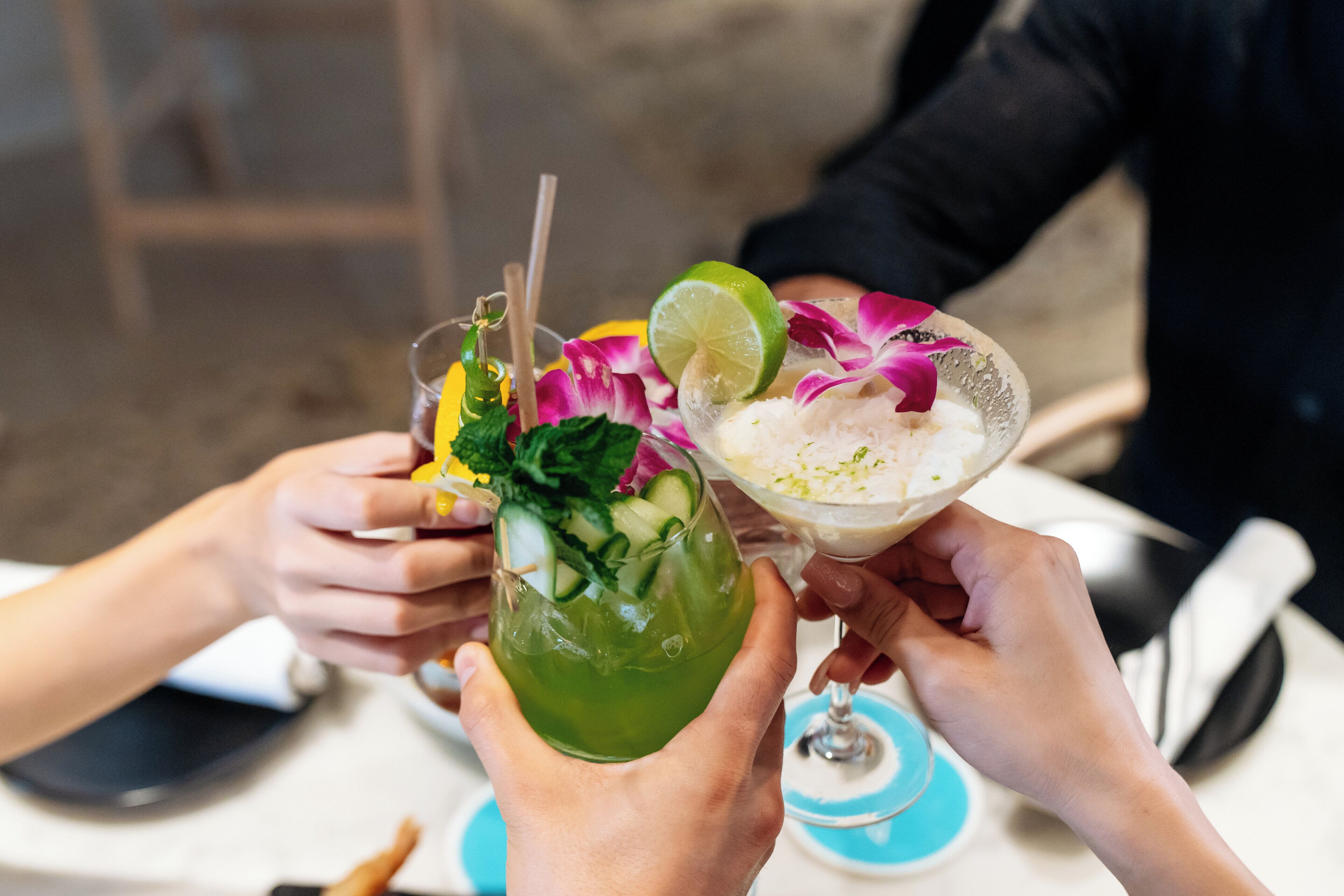 Deux cocktails décorés de quartiers de citron vert, de feuilles de menthe et de pétales de fleurs, tenus par des personnes assises à une table.