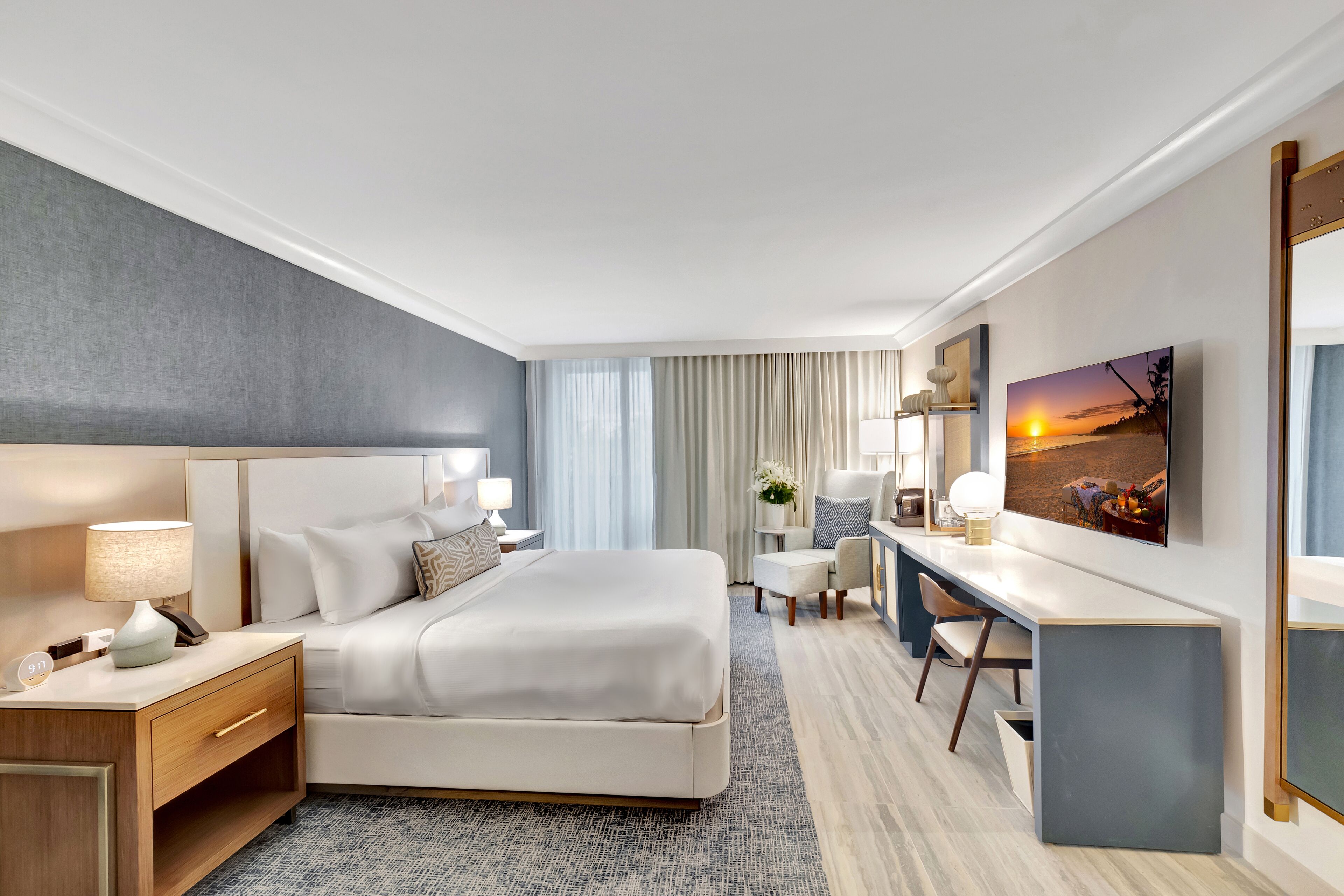 A modern hotel room with a large bed, a desk, a chair, a TV, and a painting on the wall.