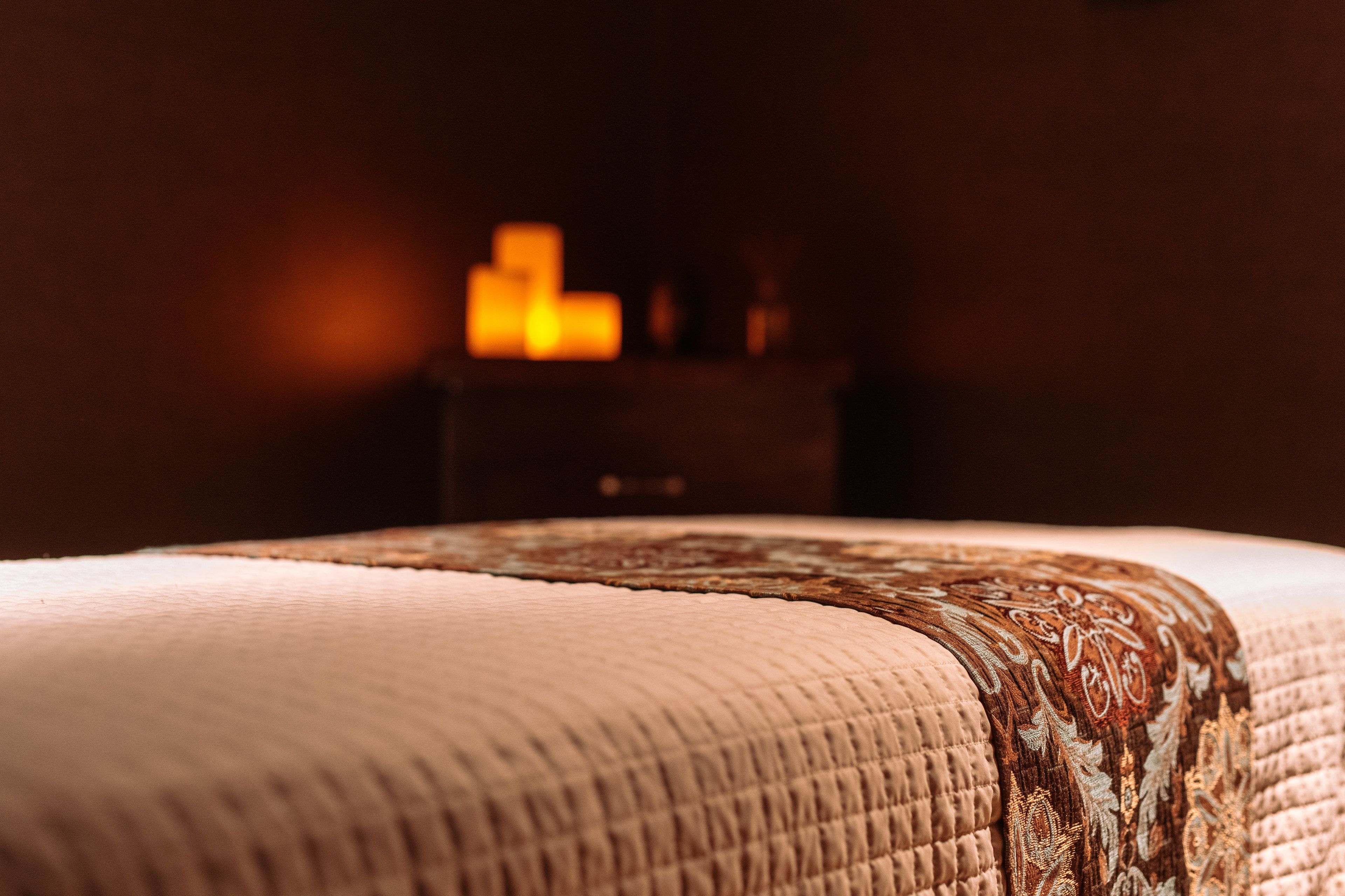 A close-up of a bed with a patterned bedspread and a dark wooden nightstand in the background.