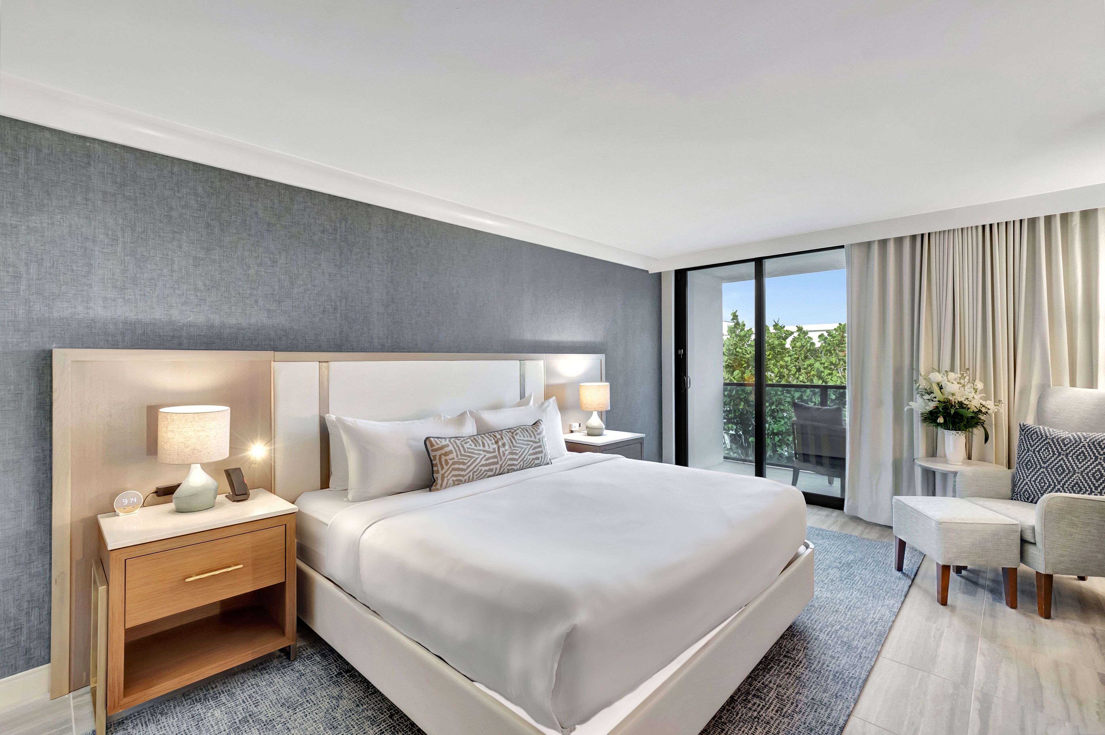 Una habitación de hotel moderna con una cama grande, mesitas de noche con lámparas, un pequeño escritorio y vistas al exterior.