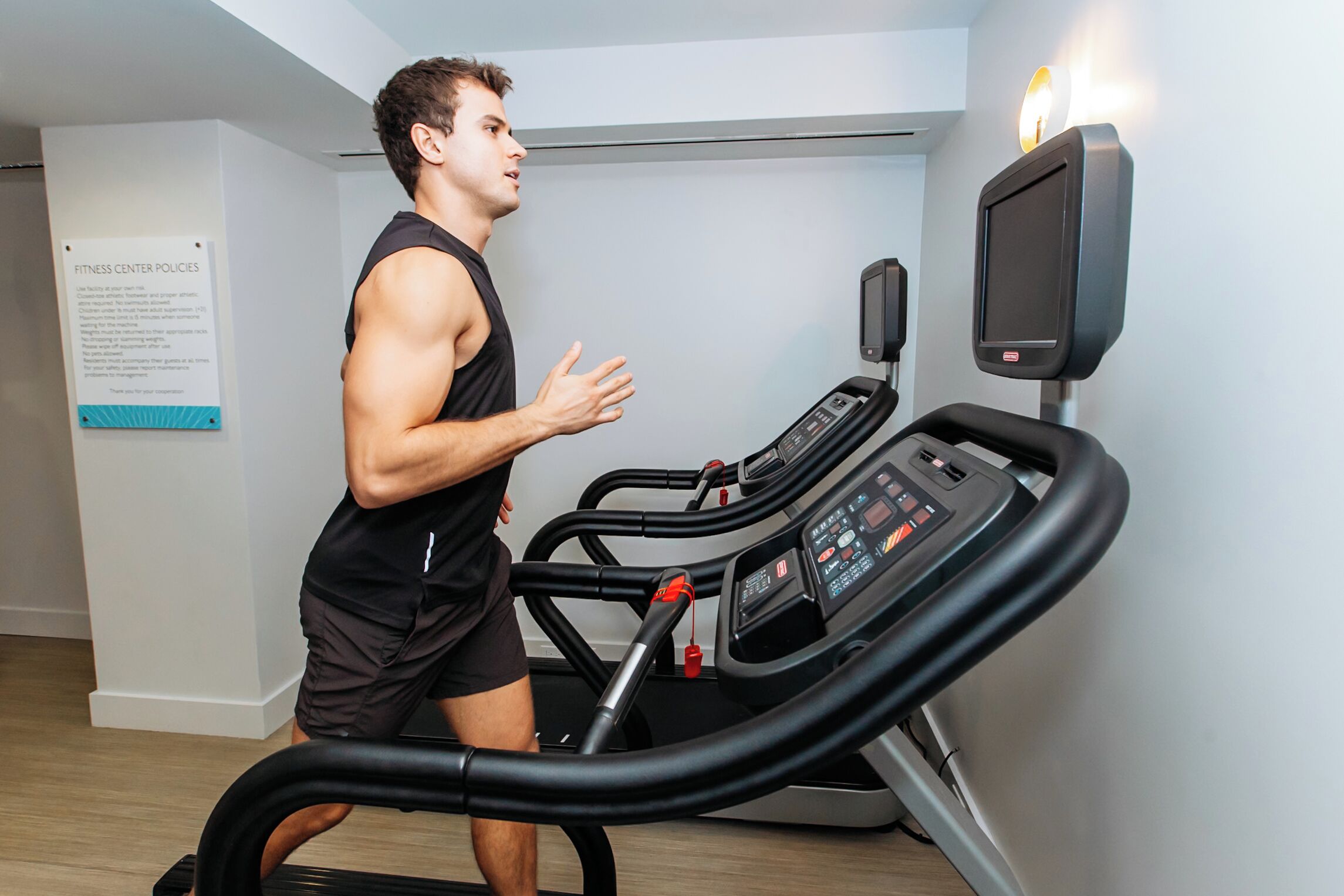 A man running on a treadmill in a room with a fitness center policy sign on the wall.