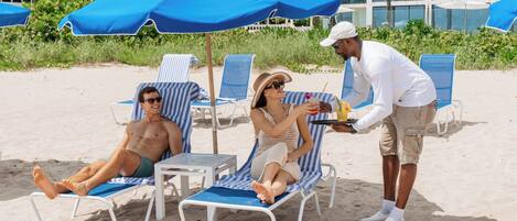 A beach setting with lounge chairs and umbrellas, a waiter serving drinks, and a building in the background.