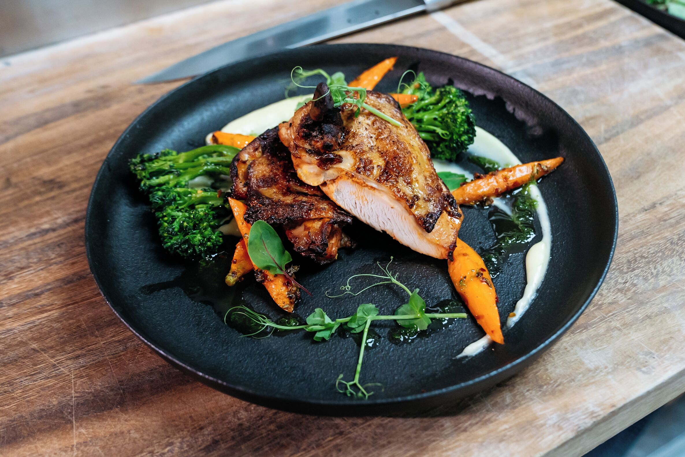A plate of food with roasted meat, vegetables, and a dark plate on a wooden surface.