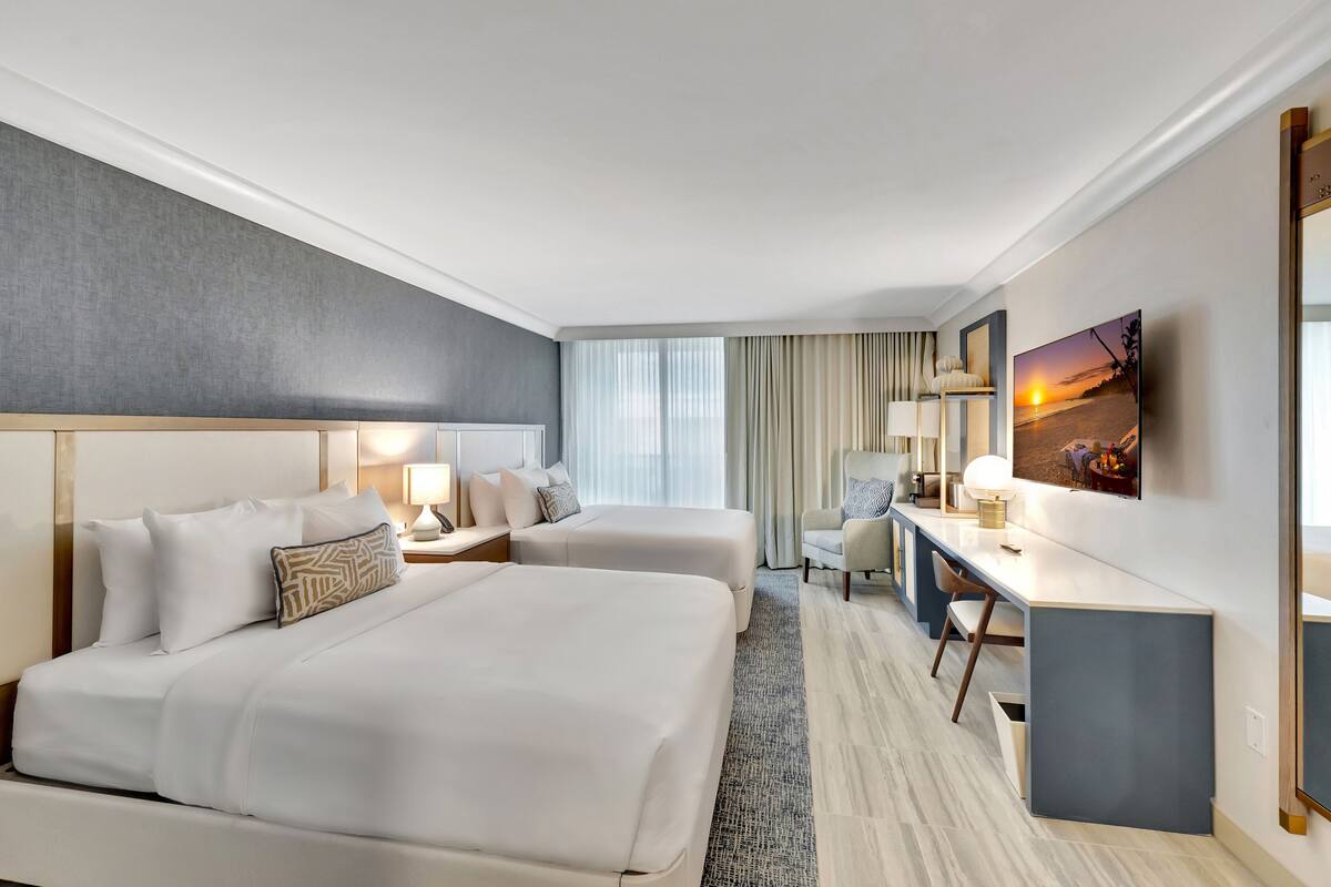 A hotel room with two beds, a desk, a chair, a TV, and a large window with curtains.