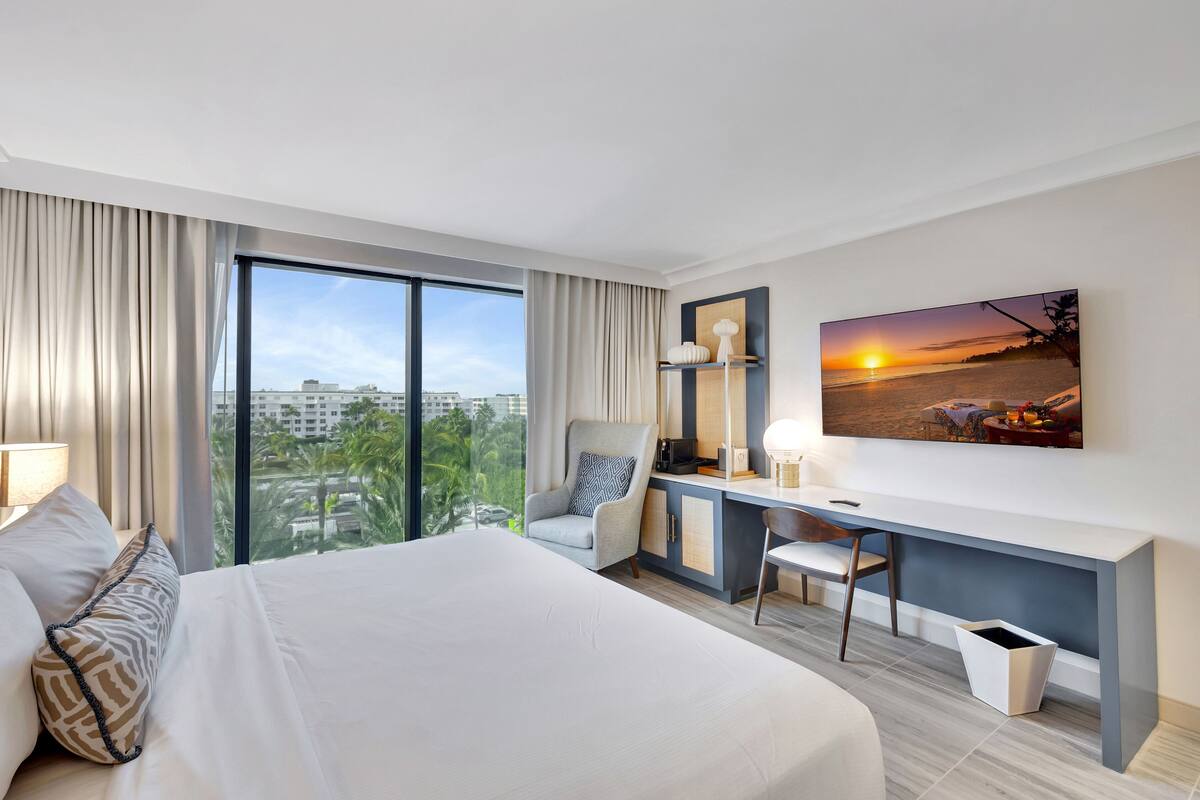 A hotel room with a large bed, a desk, a chair, a TV, and a view of the city.