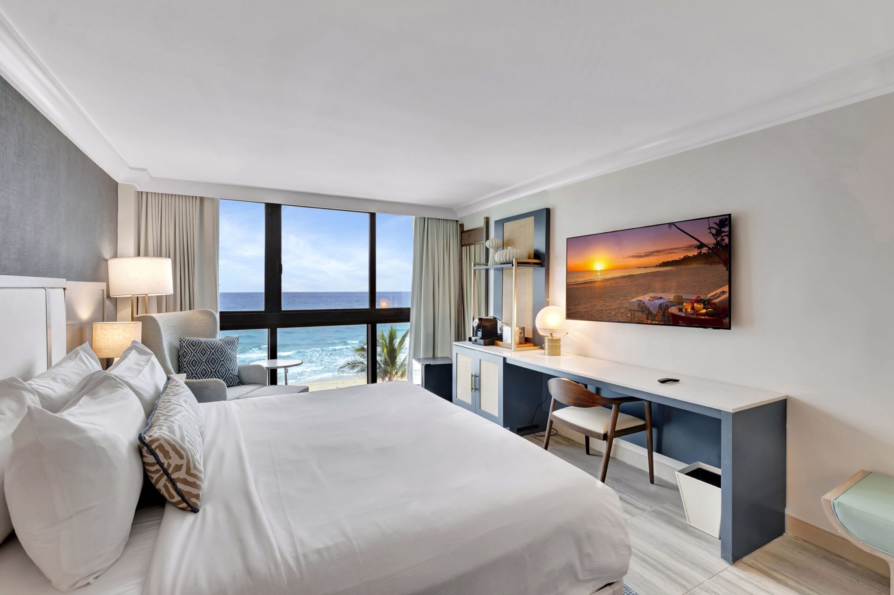 A hotel room with a large bed, a desk, a chair, a TV, and a view of the ocean.