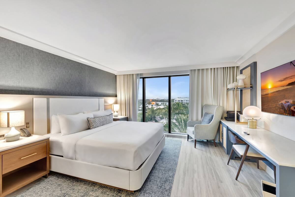 A modern hotel room with a large bed, a desk, a chair, and a view of the city.