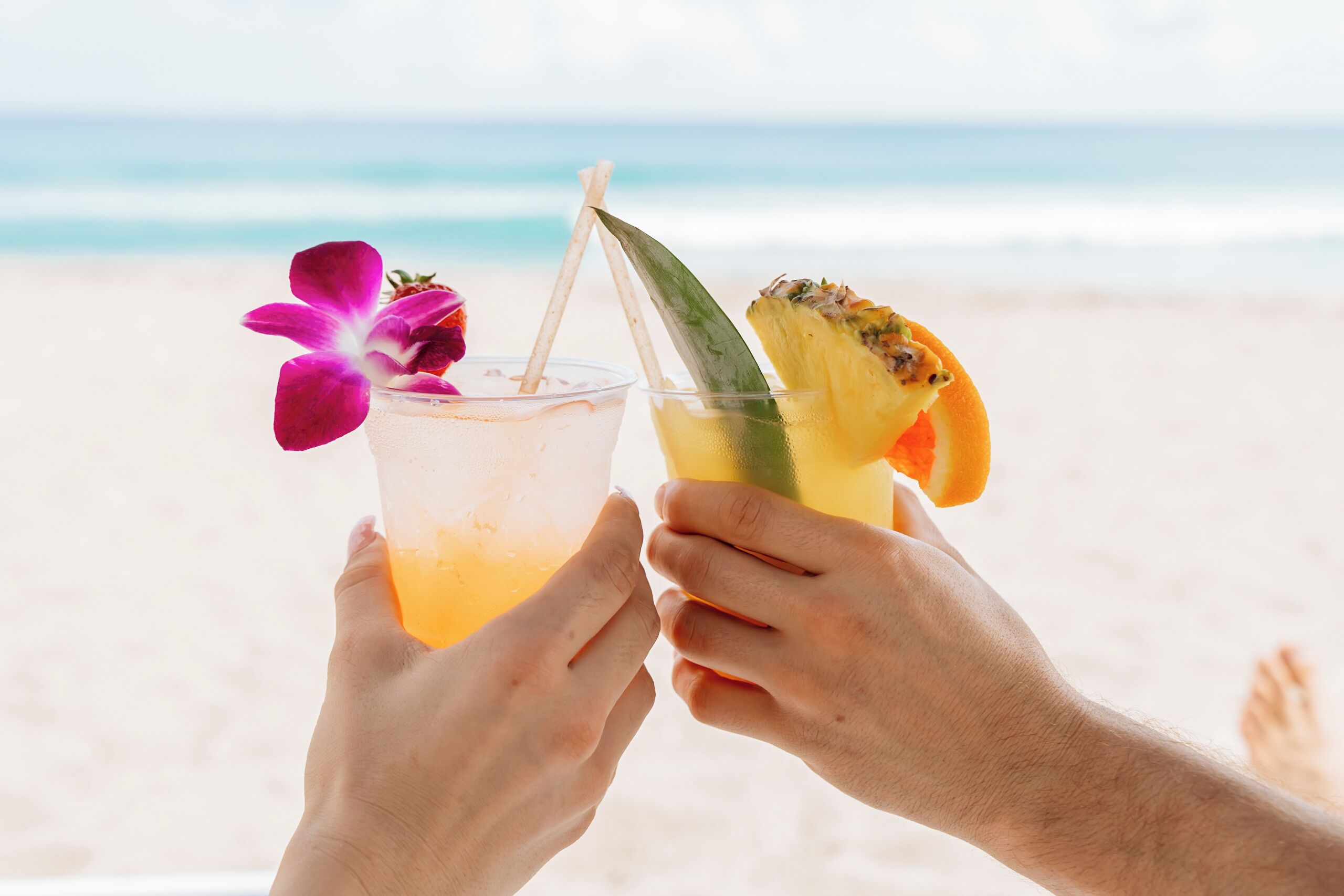 Två händer håller i glas med tropiska drinkar, med en strand och ett hav i bakgrunden.