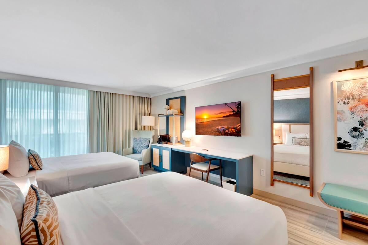 A hotel room with two beds, a desk, a chair, a desk lamp, a mirror, and a painting of a sunset.
