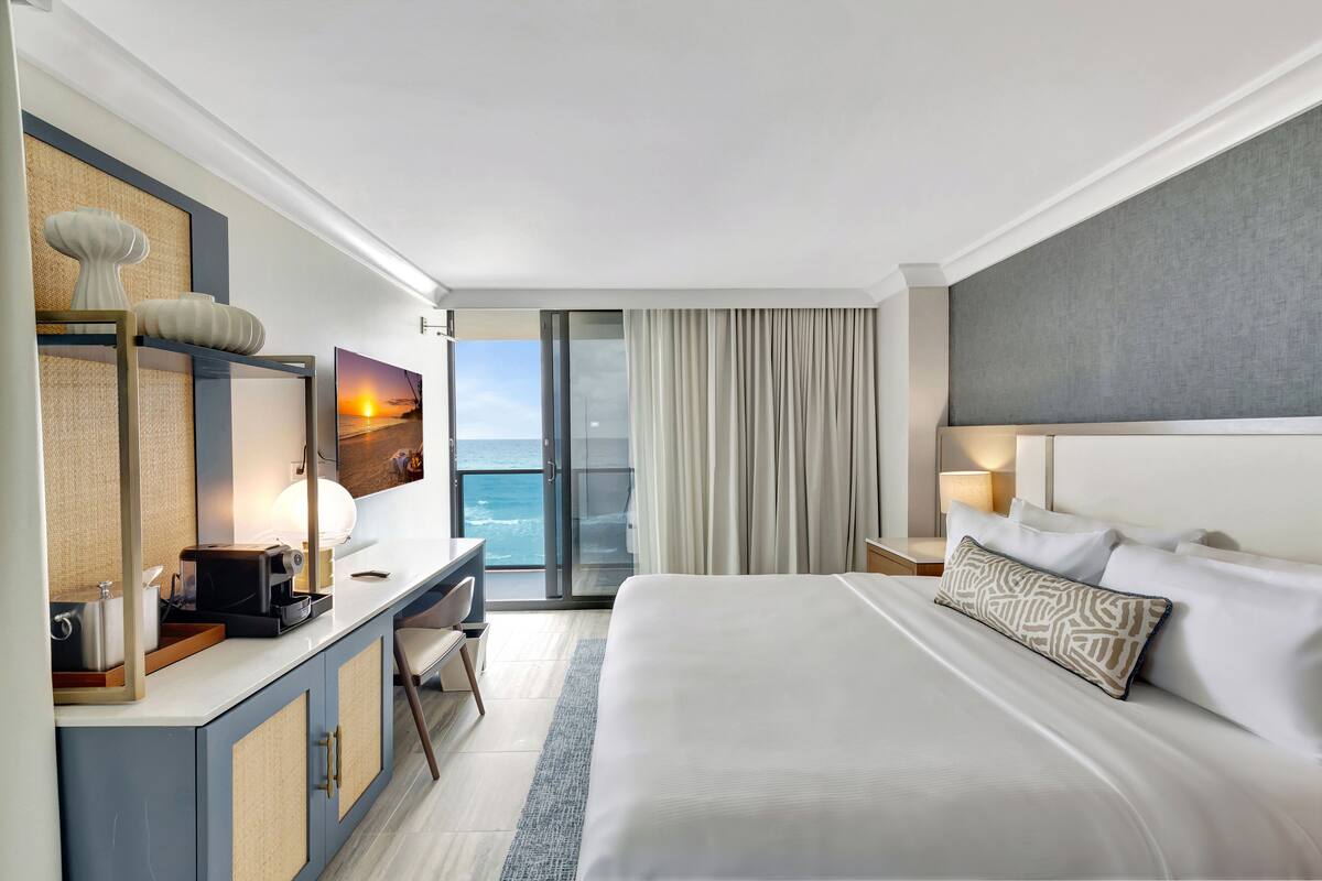 A hotel room with a large bed, a desk with a lamp, a view of the ocean through a sliding door, and a painting of a sunset.