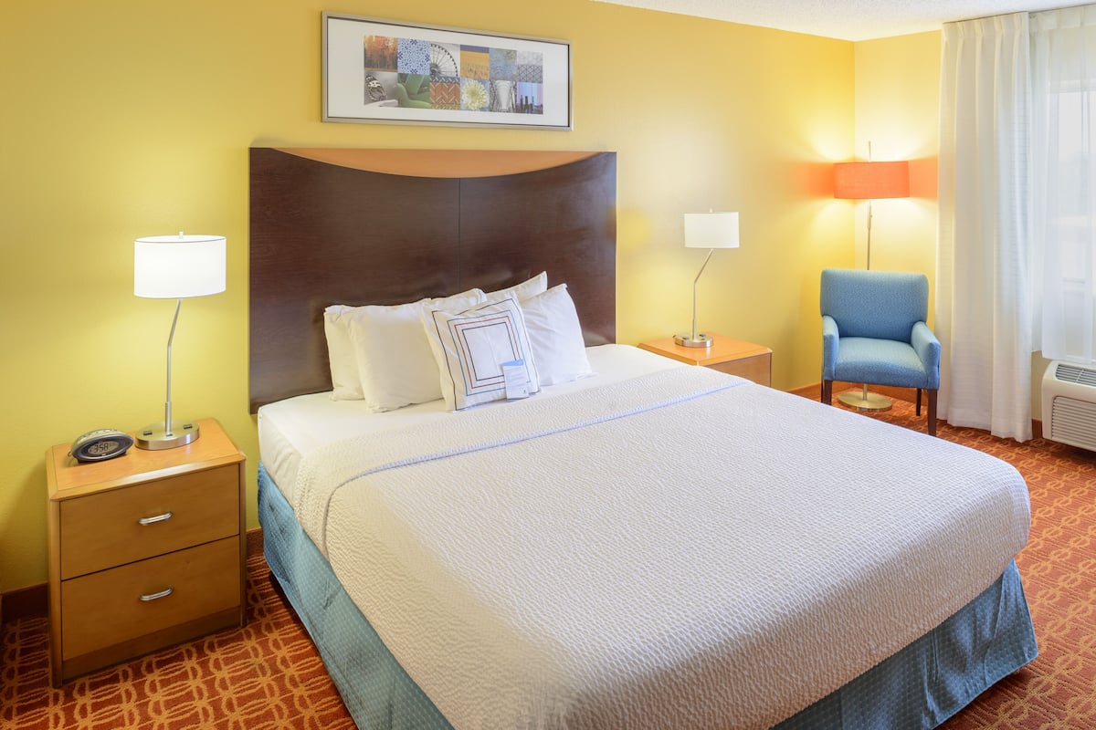 Une chambre d’hôtel avec un grand lit, une table de chevet avec une lampe, une chaise et une œuvre d’art accrochée au mur.