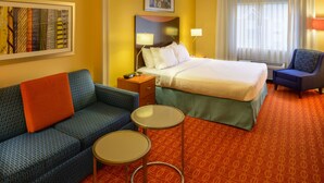 Habitación de hotel con cama, sofá, dos mesitas, una silla y un cuadro en la pared.