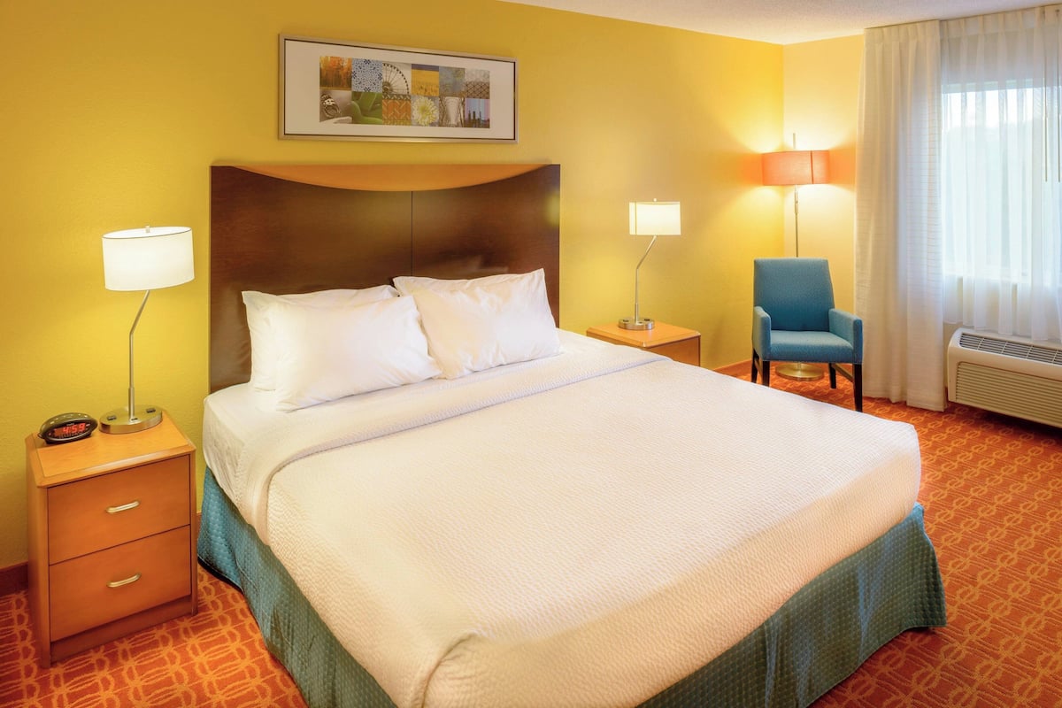 Une chambre d’hôtel avec un grand lit, deux tables de chevet, une chaise, une œuvre d’art accrochée au mur et une fenêtre avec des rideaux.
