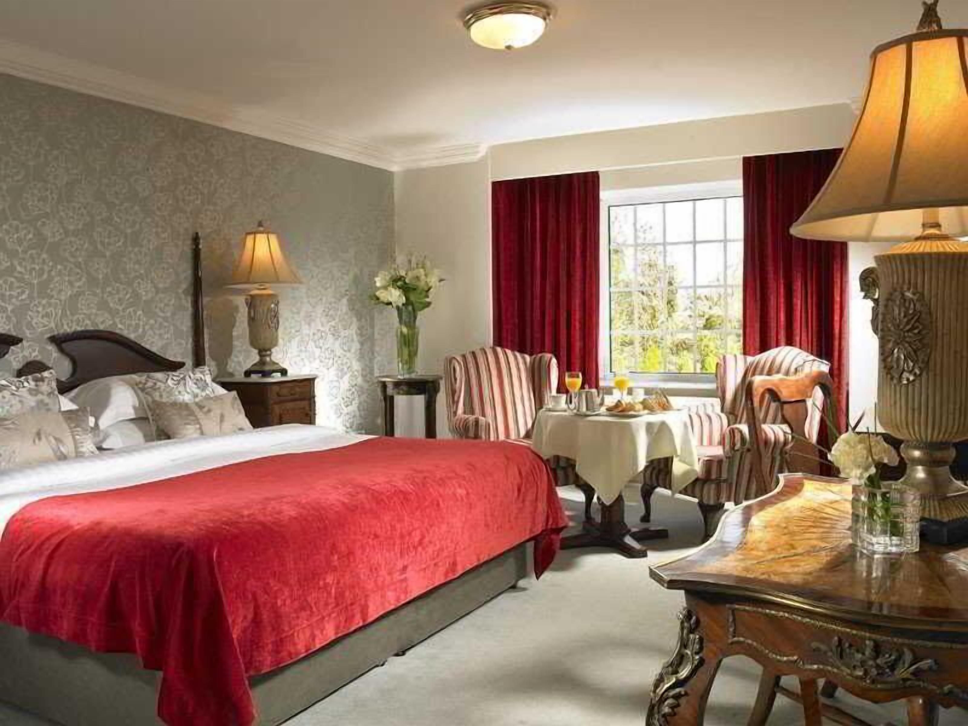 Ein Hotelzimmer mit einem großen Bett, einem kleinen Tisch mit einer Lampe, einem Sessel und einem Fenster mit Vorhängen.