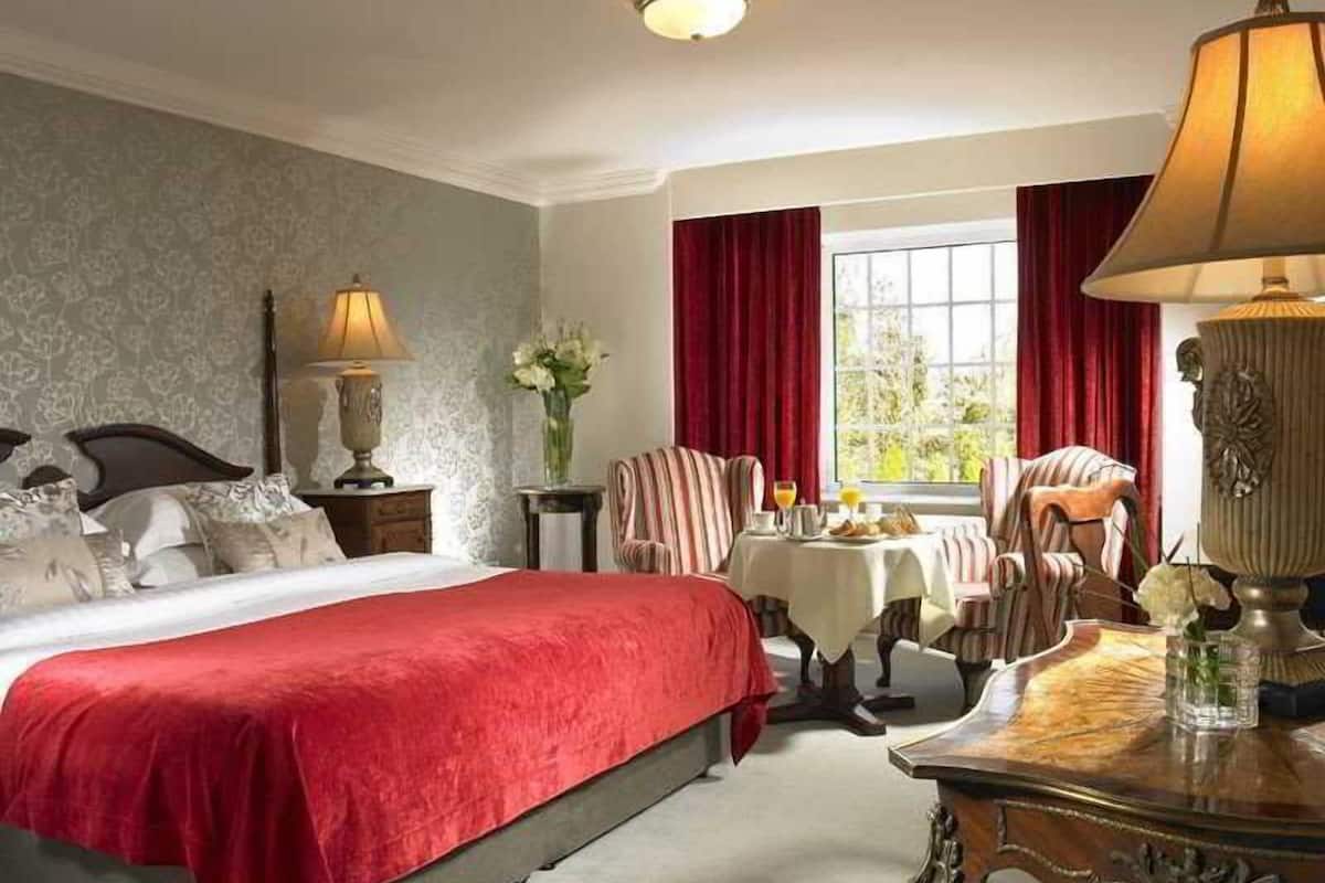 Habitación de hotel con una cama grande, una mesita con lámpara, una silla y una ventana con cortinas.